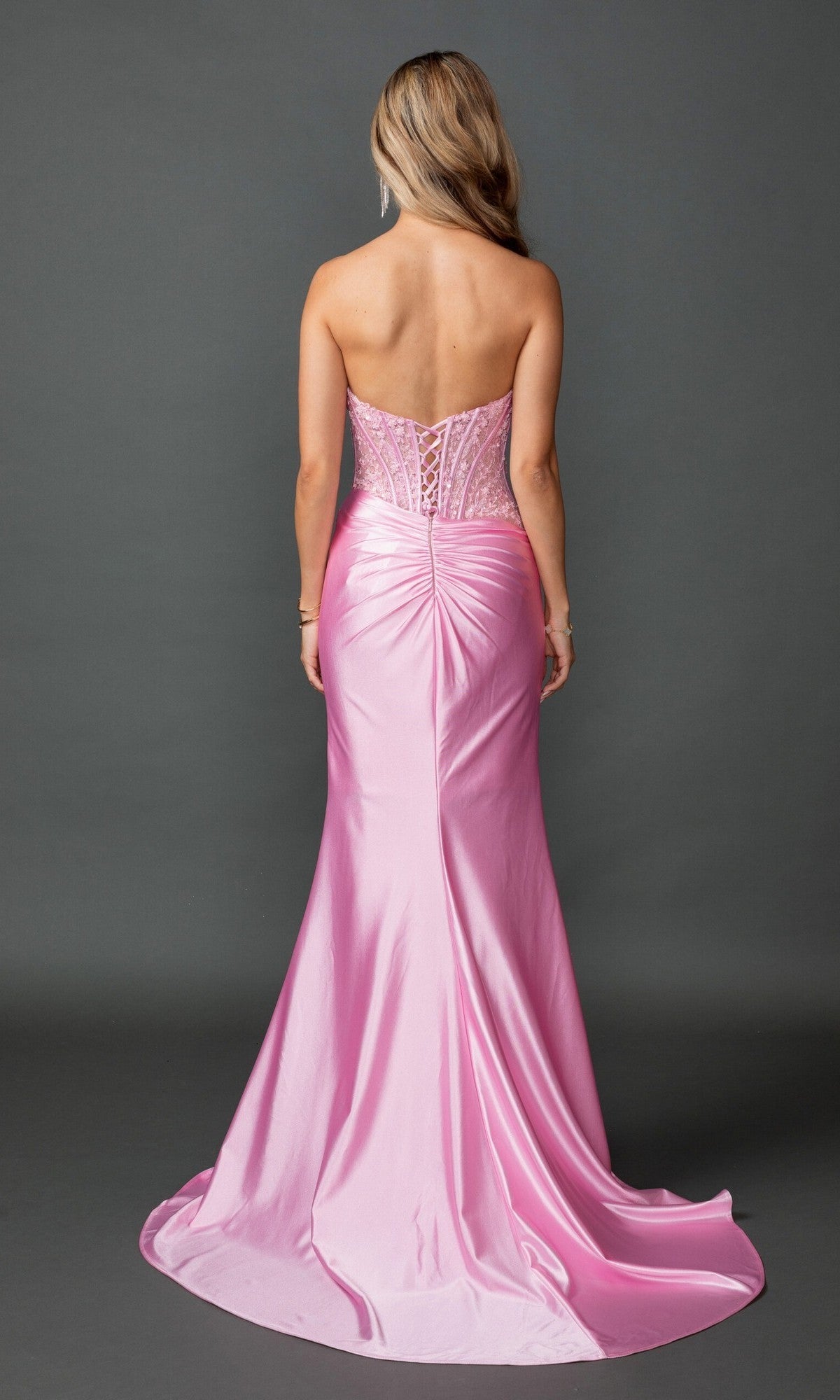 A person wearing the Long Prom Dress: Dancing Queen 4587, a pink strapless gown featuring a satin fitted sheath skirt, corset bodice, ruched back, and slight train, stands against a plain gray background.