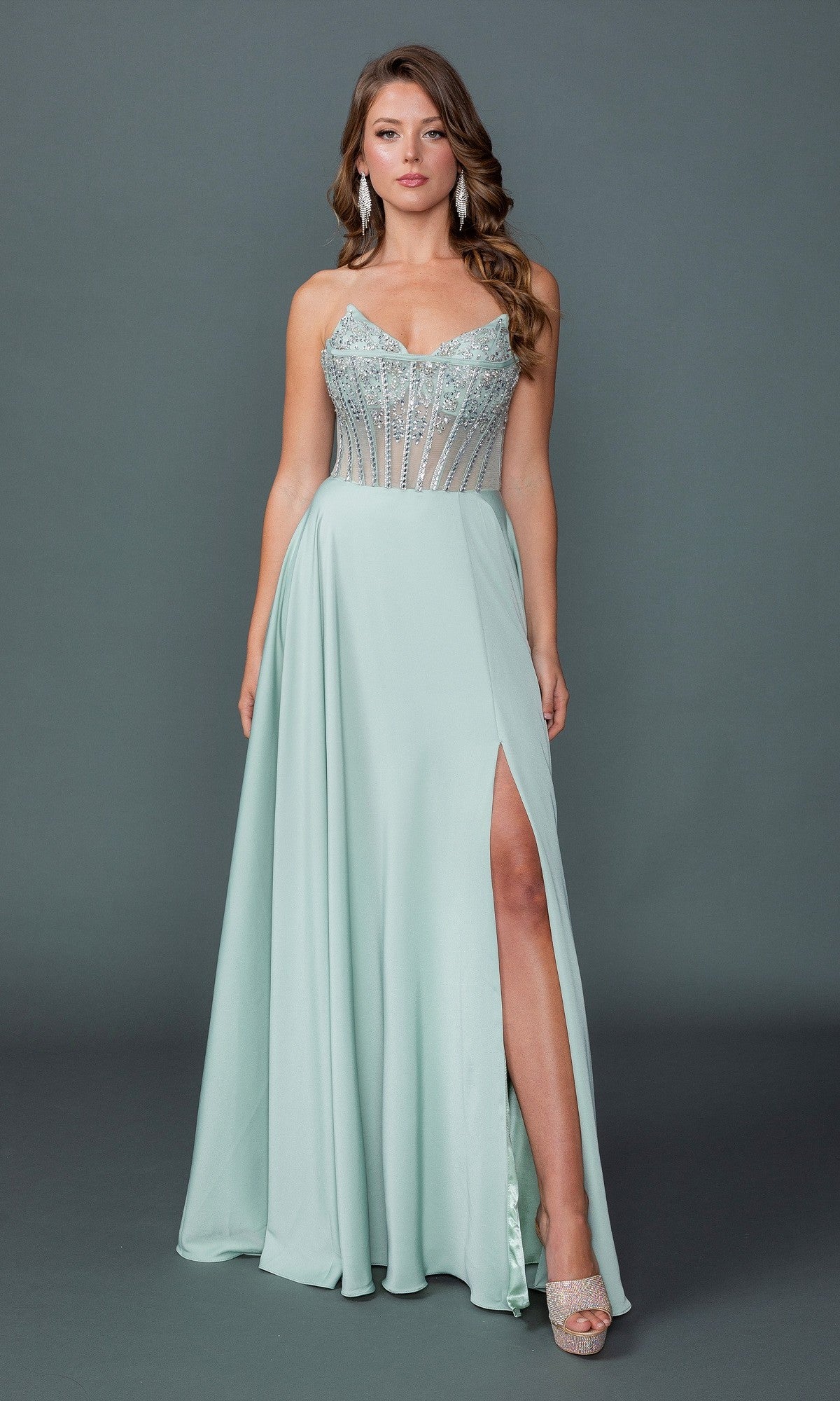 A woman in a sage Long Prom Dress: Dancing Queen 4589 with a beaded corset bodice and thigh-high slit pairs it with silver heels, posing against a plain gray background.