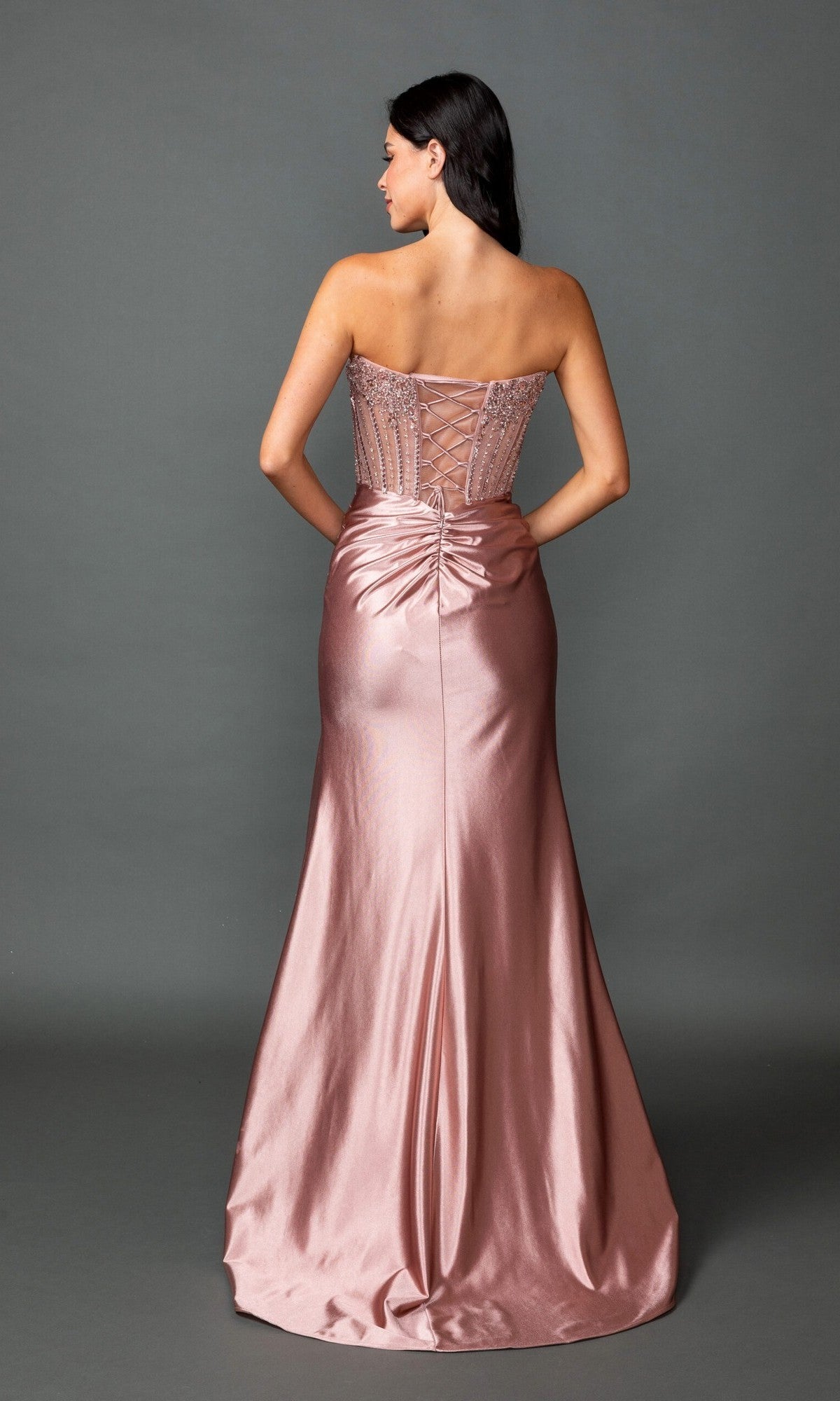 A woman stands facing away, wearing the Long Prom Dress: Dancing Queen 4592—a strapless pink gown with a fitted corset bodice, satin sheath skirt, lace-up back, and ruched detailing—set against a plain gray backdrop.