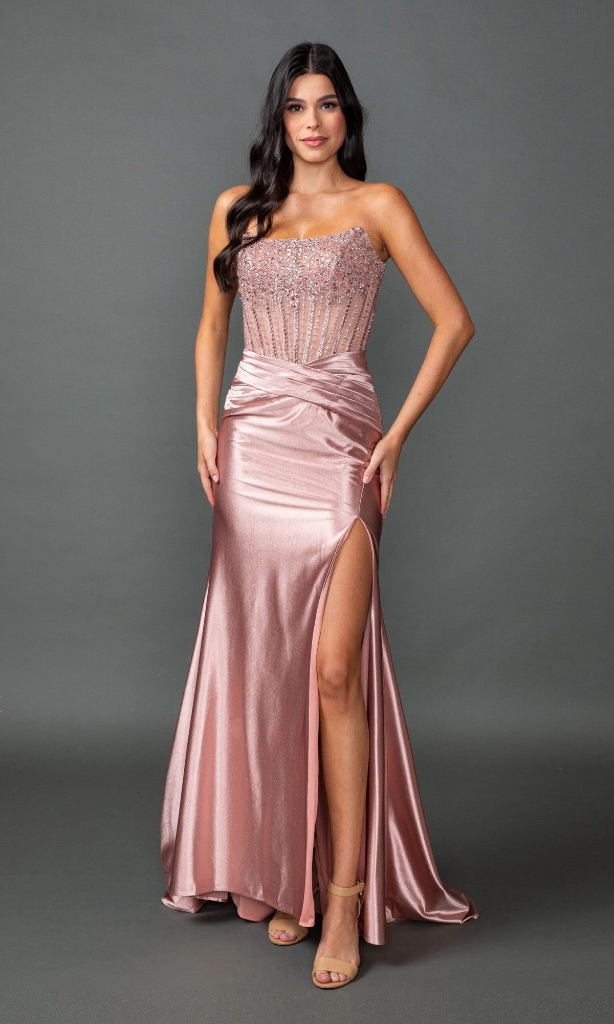 A woman wearing the Long Prom Dress: Dancing Queen 4592 in rose gold, which features a strapless beaded corset bodice and a satin sheath skirt with a high slit, stands against a plain gray background.