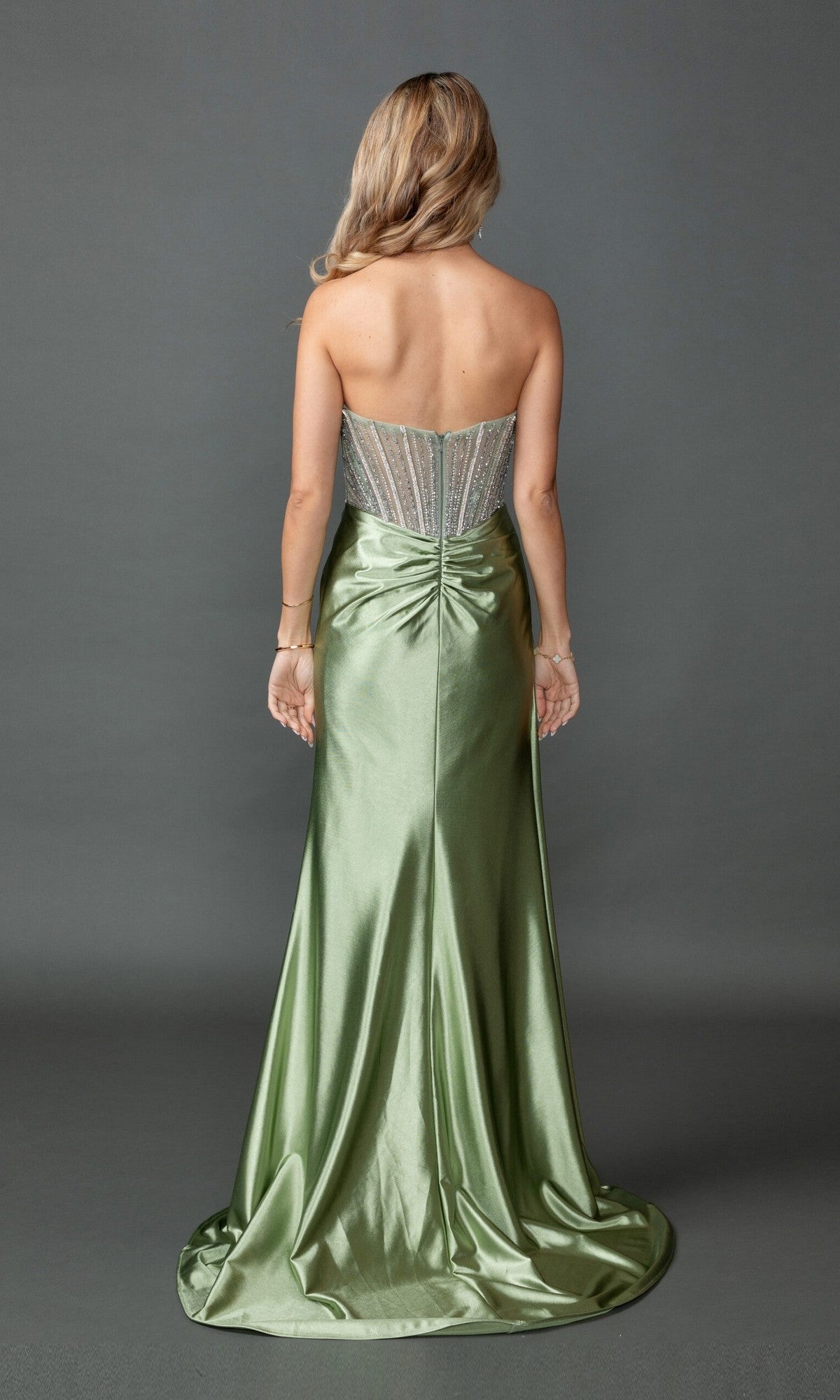 A woman stands facing away on a gray backdrop, wearing the Long Prom Dress: Dancing Queen 4593, which features a beaded, sheer corset bodice and a flowing green satin skirt.