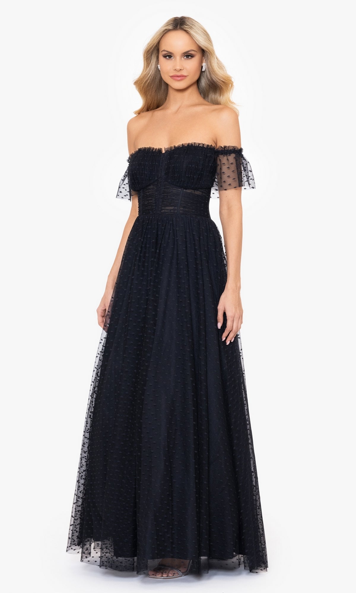 A woman wears the Swiss Dot Long Black Lace-Up Prom Dress 4601BN with an off-the-shoulder design, sheer Swiss dot overlay, and ruffled sleeves, standing against a plain white background.