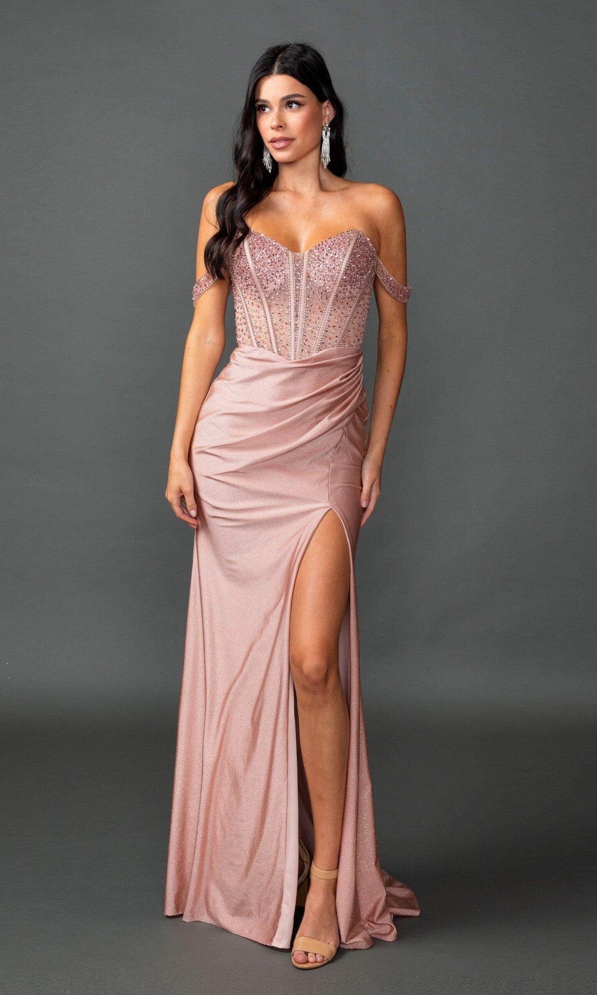A woman with long dark hair and drop earrings stands against a gray background wearing the Dancing Queen 4601 long prom dress in rose gold, featuring an off-the-shoulder neckline, sheer corset bodice, thigh-high slit, and strappy sandals.