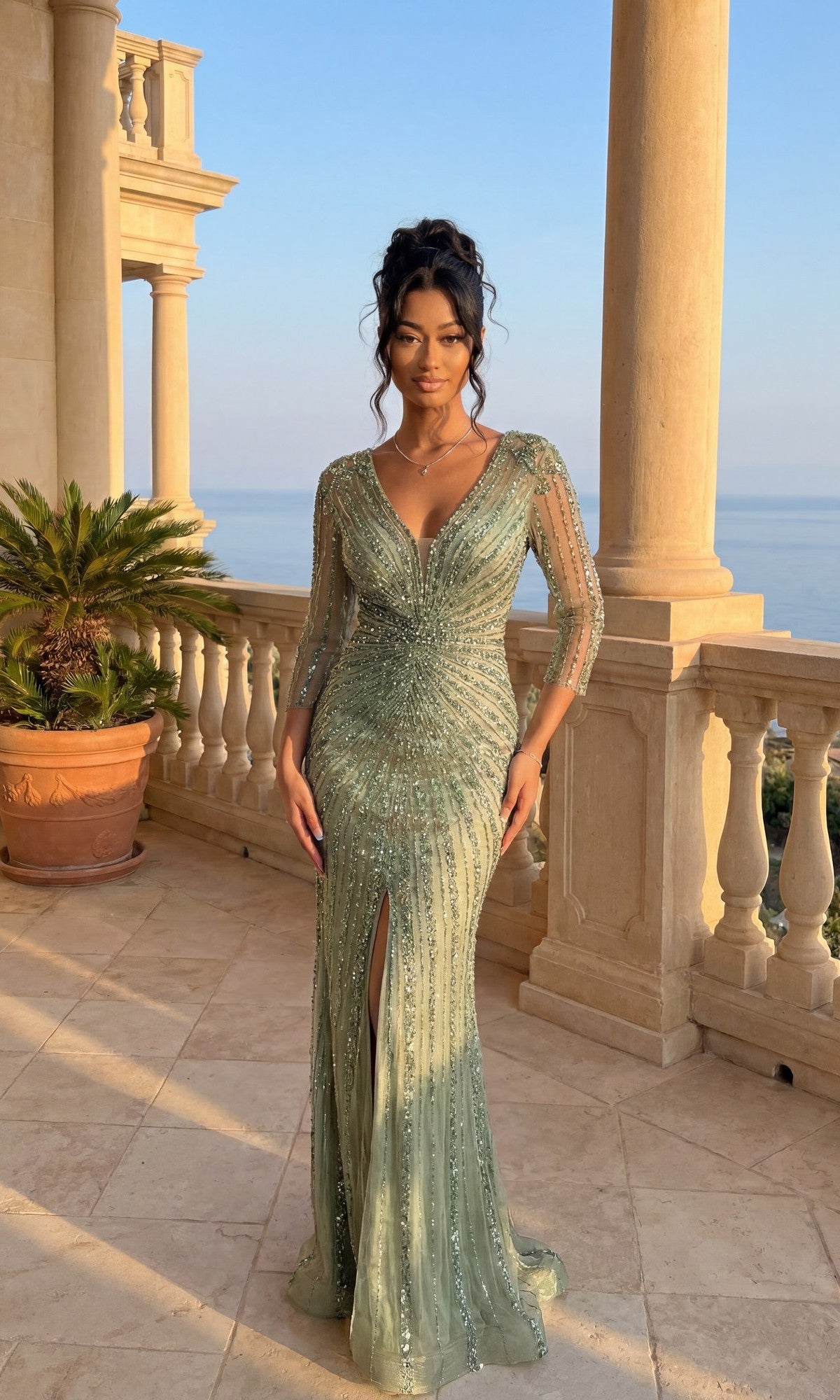 A woman in the olive Dancing Queen 4612 long formal dress, featuring beading, sheer sleeves, and a front leg slit, stands on a stone balcony overlooking the sea with columns and a potted palm behind her.