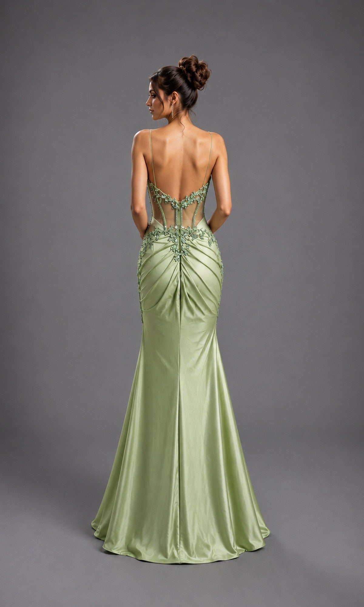 A woman wearing the Long Prom Dress: Dancing Queen 4614, a fitted sage green satin gown with beaded details, an open back, and a sleek corset bodice, stands against a plain gray background.