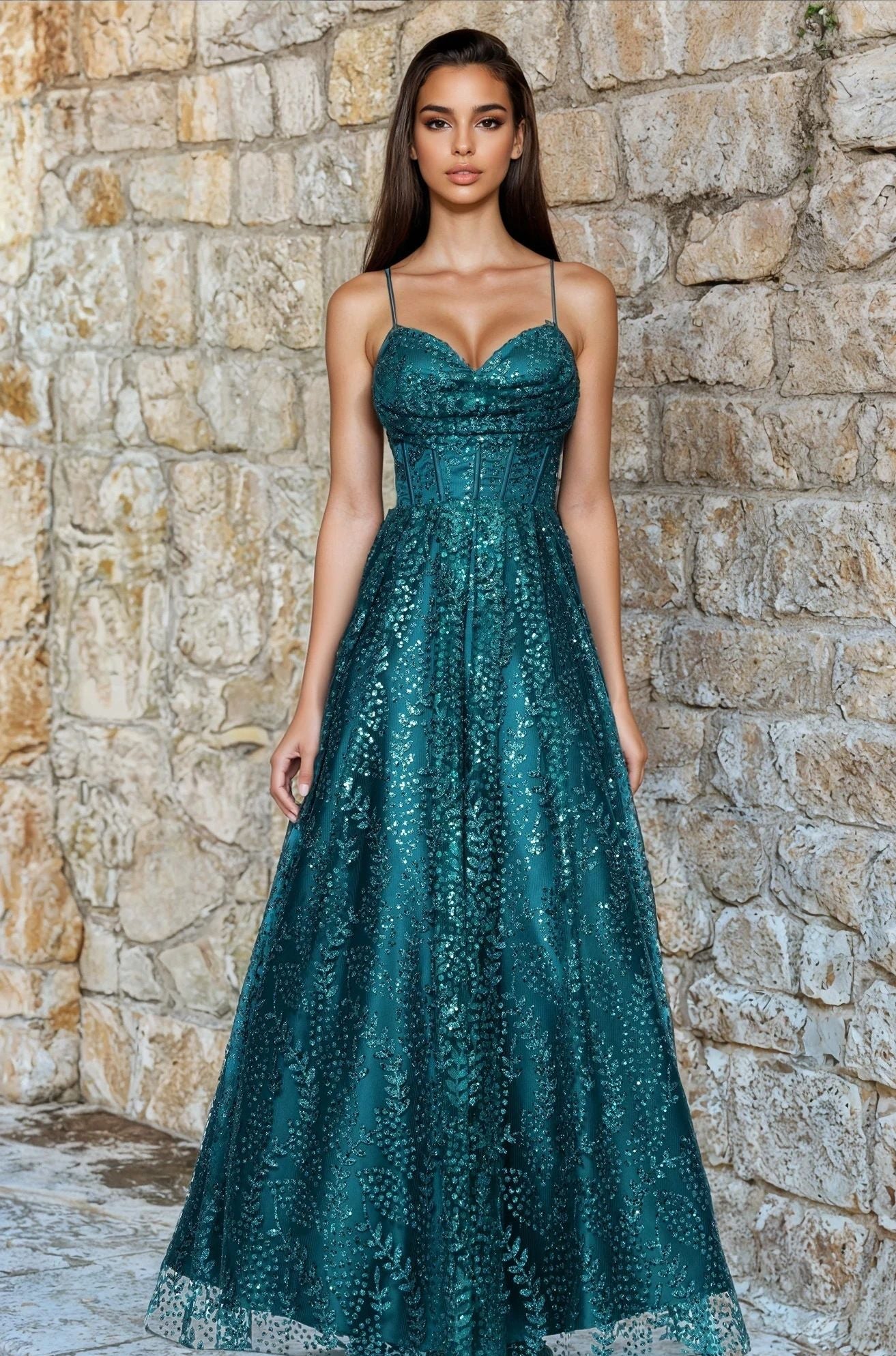 A woman stands before a stone wall wearing the Blondie Nites 4617BN Long Glitter Prom Dress in teal, featuring thin straps and a fitted, embellished corset-style bodice with an A-line, floor-length silhouette.