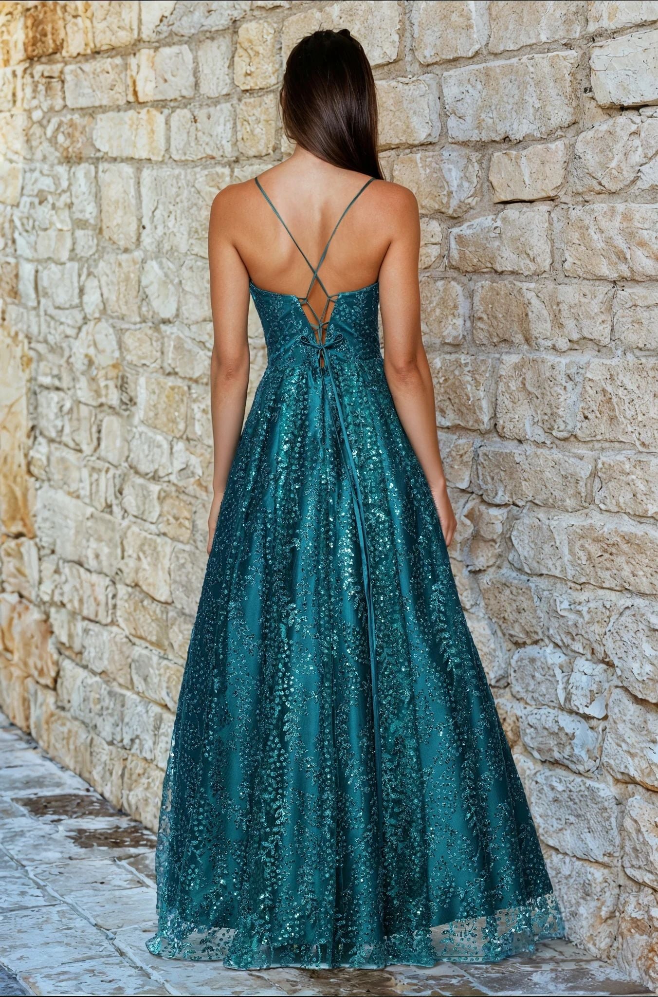 A woman faces away from the camera against a stone wall, wearing the Blondie Nites 4617BN long glitter prom dress in teal, featuring a floor-length A-line skirt, corset-style bodice, sequin details, and crisscross straps.