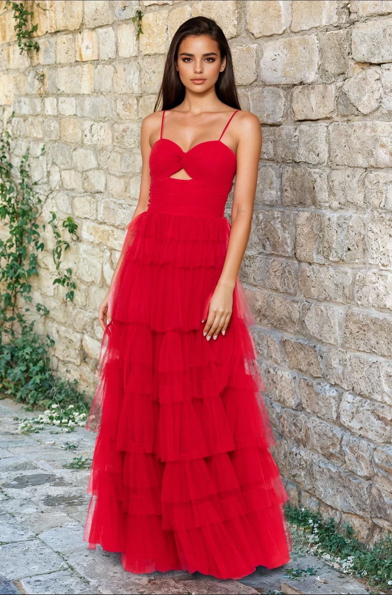 Outdoors against a stone wall, a woman wears the Blondie Nites 4637BN, a long red ruffled prom dress with a tiered tulle skirt, thin straps, sweetheart bodice, and small cutout below the bust.