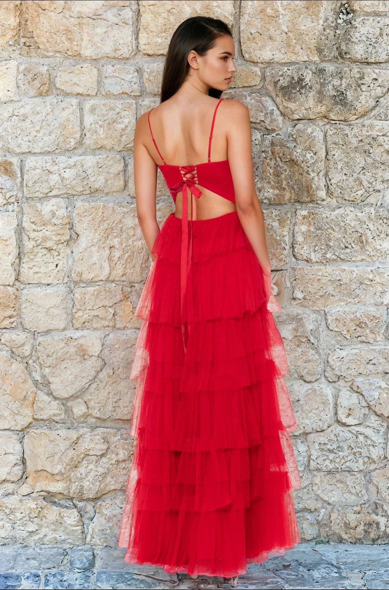 A woman poses before a stone wall in the Blondie Nites 4637BN—a sleeveless, backless long red prom dress featuring a lace-up back, thin straps, and tiered A-line ruffles.