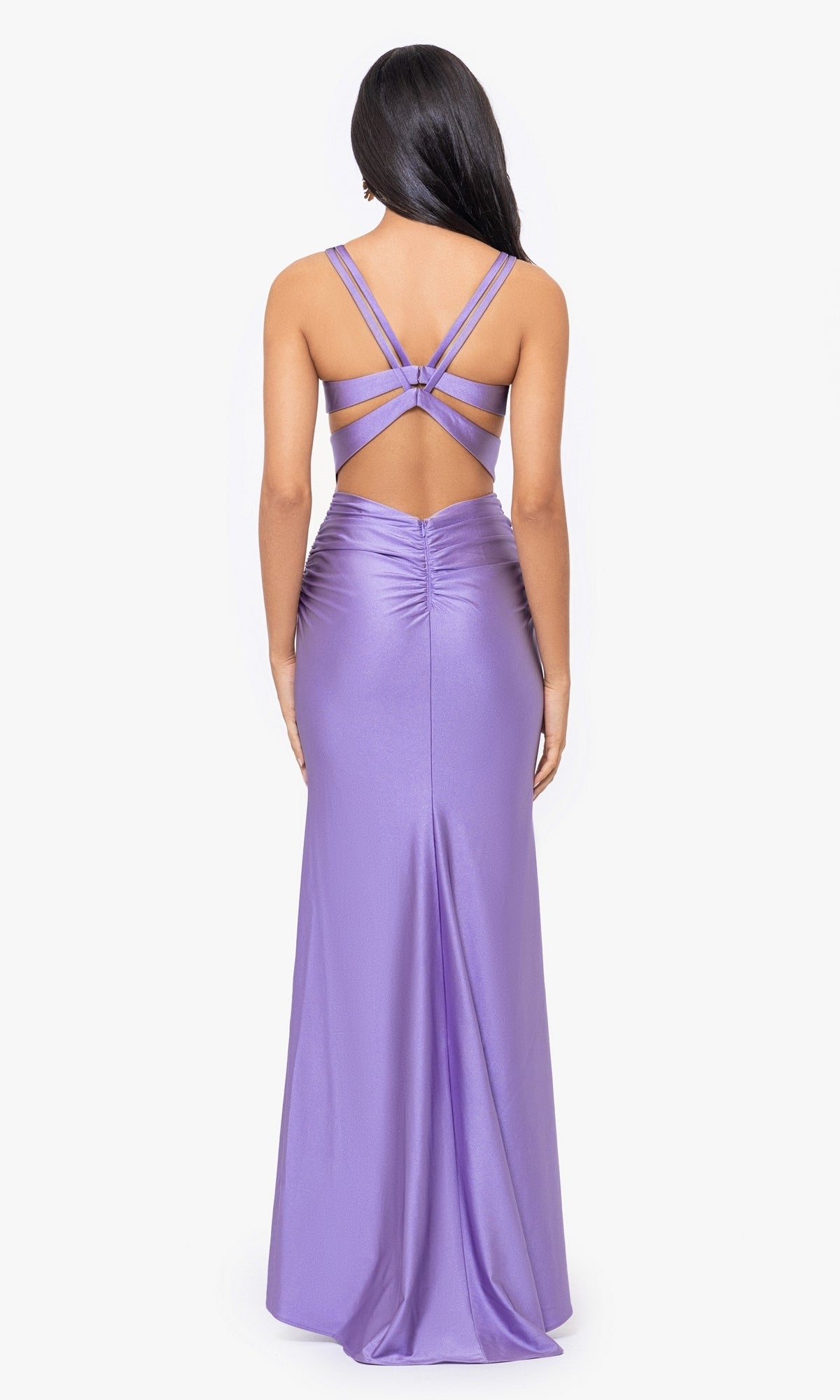 A woman is shown from behind wearing the Blondie Nites Long Lavender Prom Dress 4639BN, featuring a floor-length silhouette, ruched back, and crisscross strap detailing against a white background.