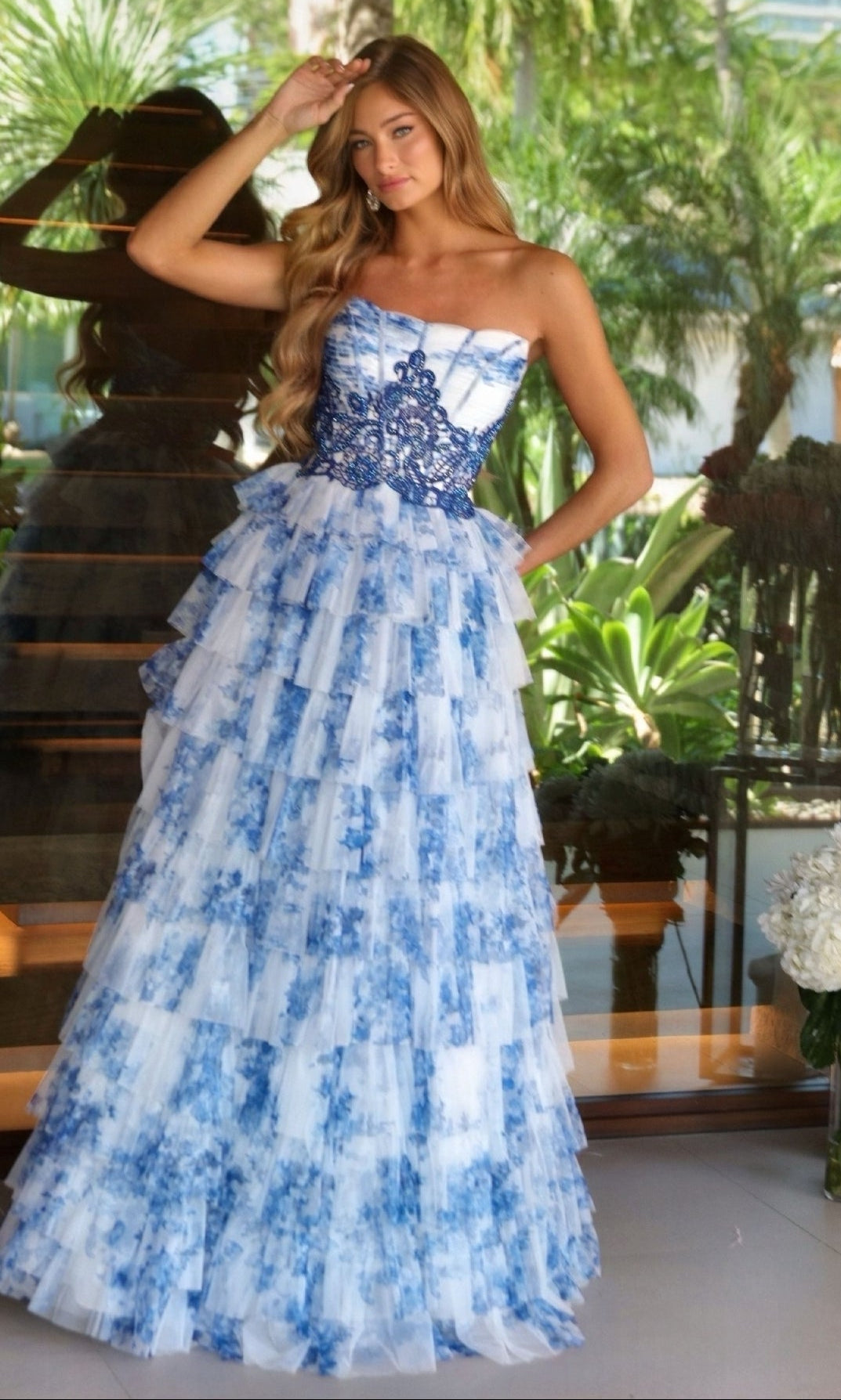 A woman stands in front of glass doors, wearing the Ava Presley 47106—a strapless white and blue floral tiered A-line prom dress—surrounded by lush greenery.