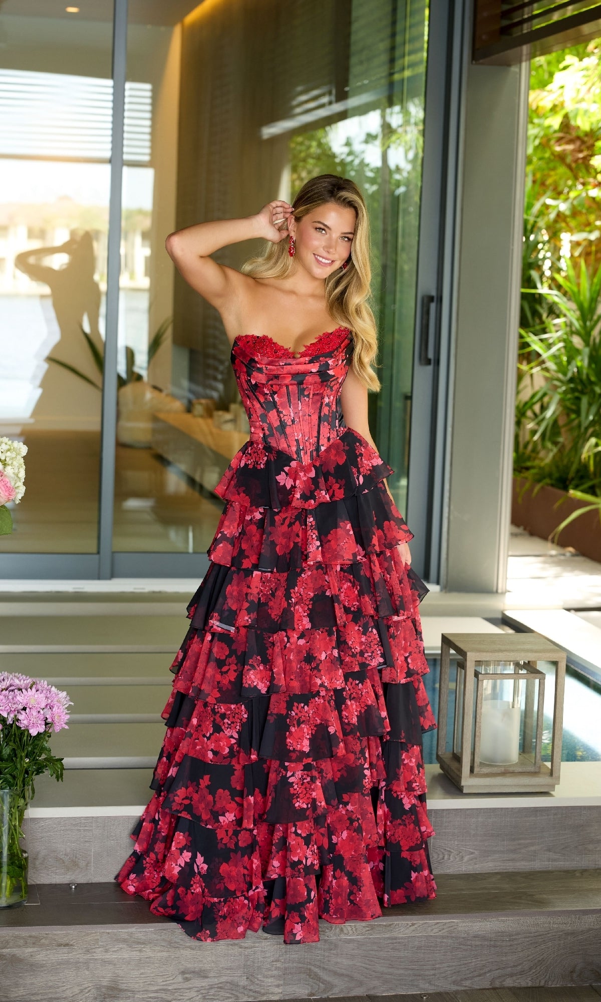 A woman wearing the Ava Presley 47108 long prom dress, a strapless tiered black and red floral gown with a corset bodice, poses outside a modern house near a lantern and a bouquet of flowers.