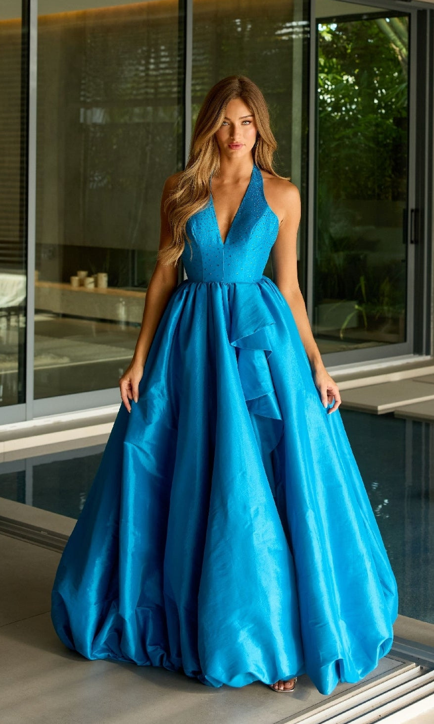 A woman in a bright blue Ava Presley 47146 Halter Long Balloon Prom Dress stands indoors near large glass windows and a pool.
