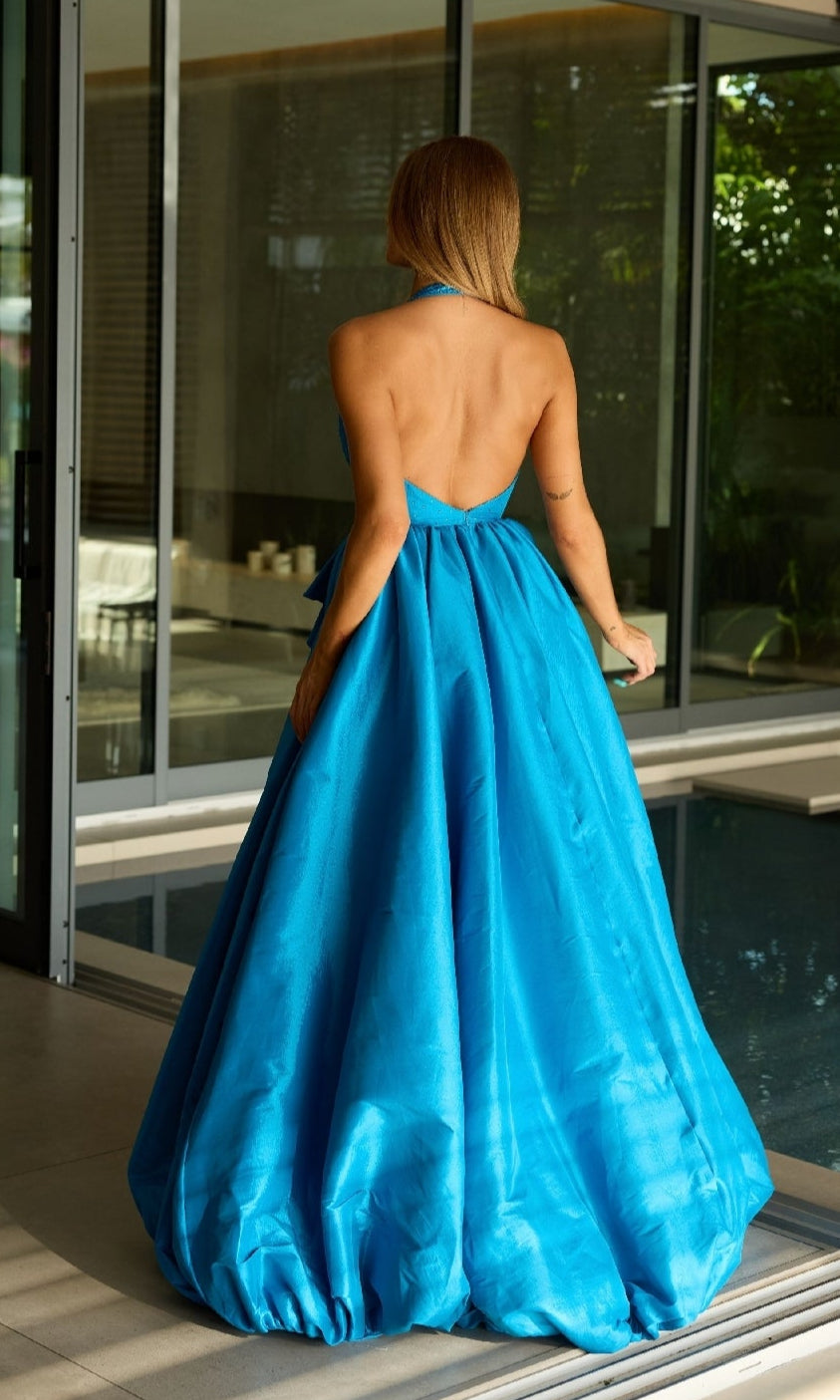 A woman stands indoors by a pool, facing away from the camera, wearing the Ava Presley 47146 Halter Long Balloon Prom Dress in bright blue.