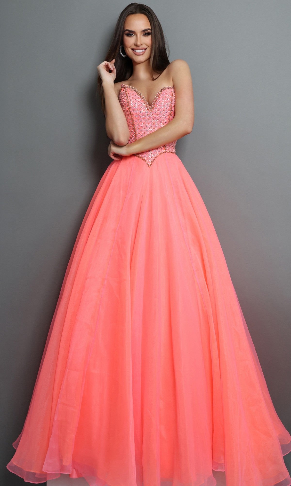 A woman is wearing the Long Formal Dress: Jovani 47196, a stunning floor-length designer gown in bright coral pink. The strapless dress features a beautifully beaded bodice and creates an elegant silhouette against a plain gray background, making it the perfect choice for special occasions.