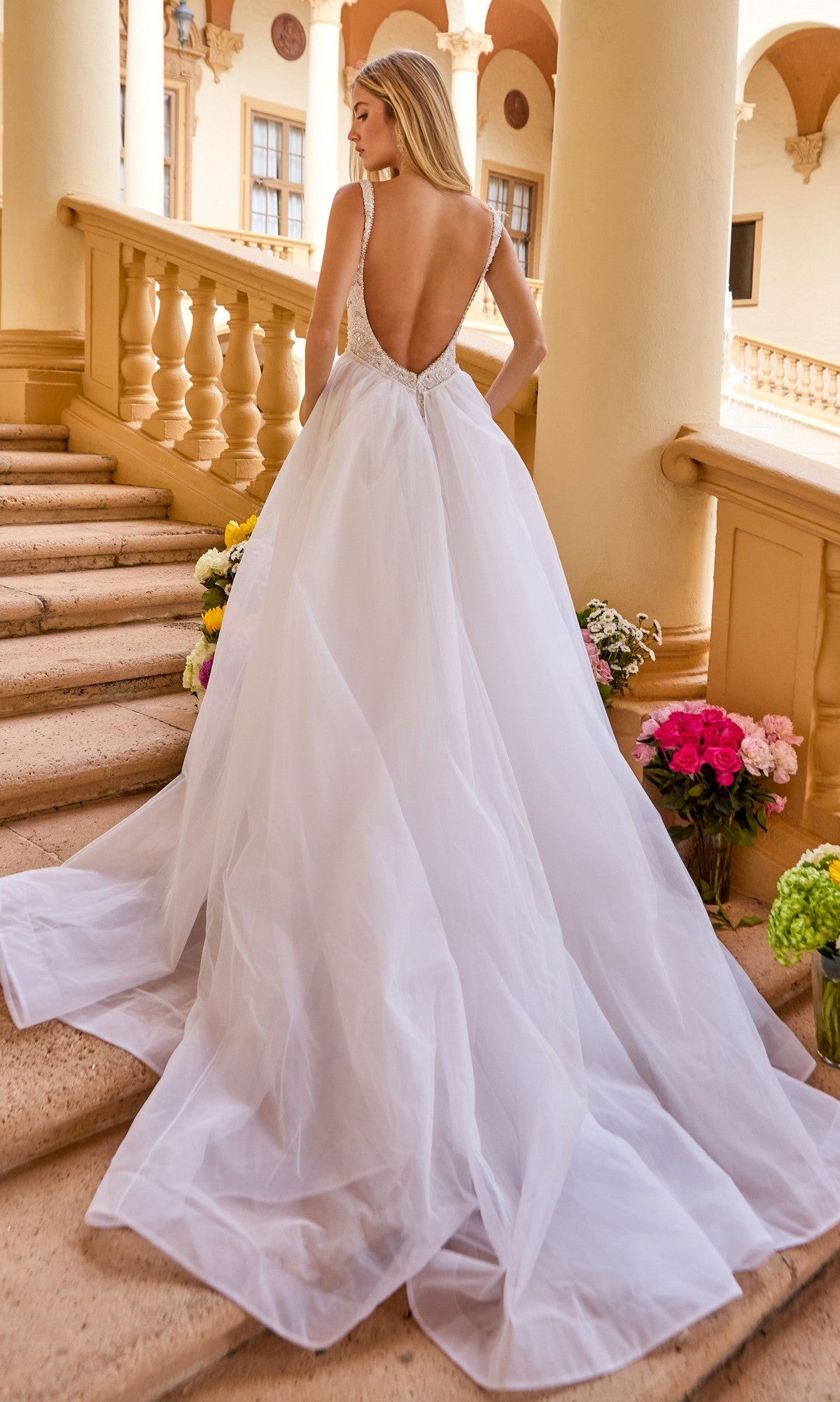 A woman in the Ava Presley 47224 long formal dress with an open back stands on stone stairs adorned with colorful flowers, facing away from the camera.