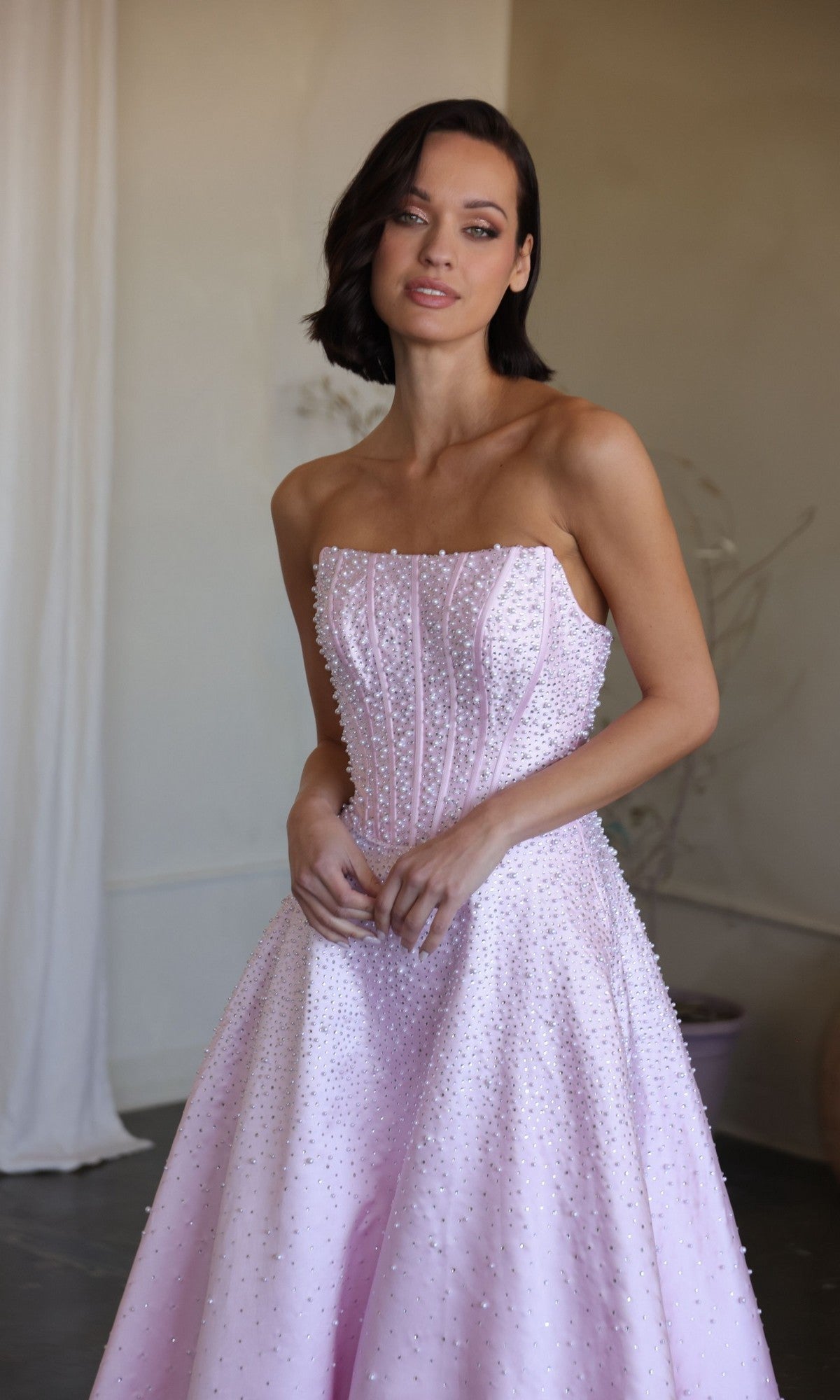 A woman in the Ava Presley 47283 long prom dress, featuring a strapless corset bodice and delicate pearl details, stands indoors in a light pink gown, gazing slightly to the side.