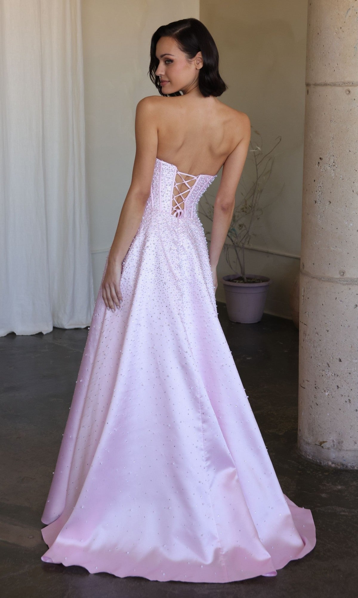 A woman stands indoors, looking over her shoulder in the Ava Presley 47283 long prom dress with a strapless corset bodice and lace-up back.