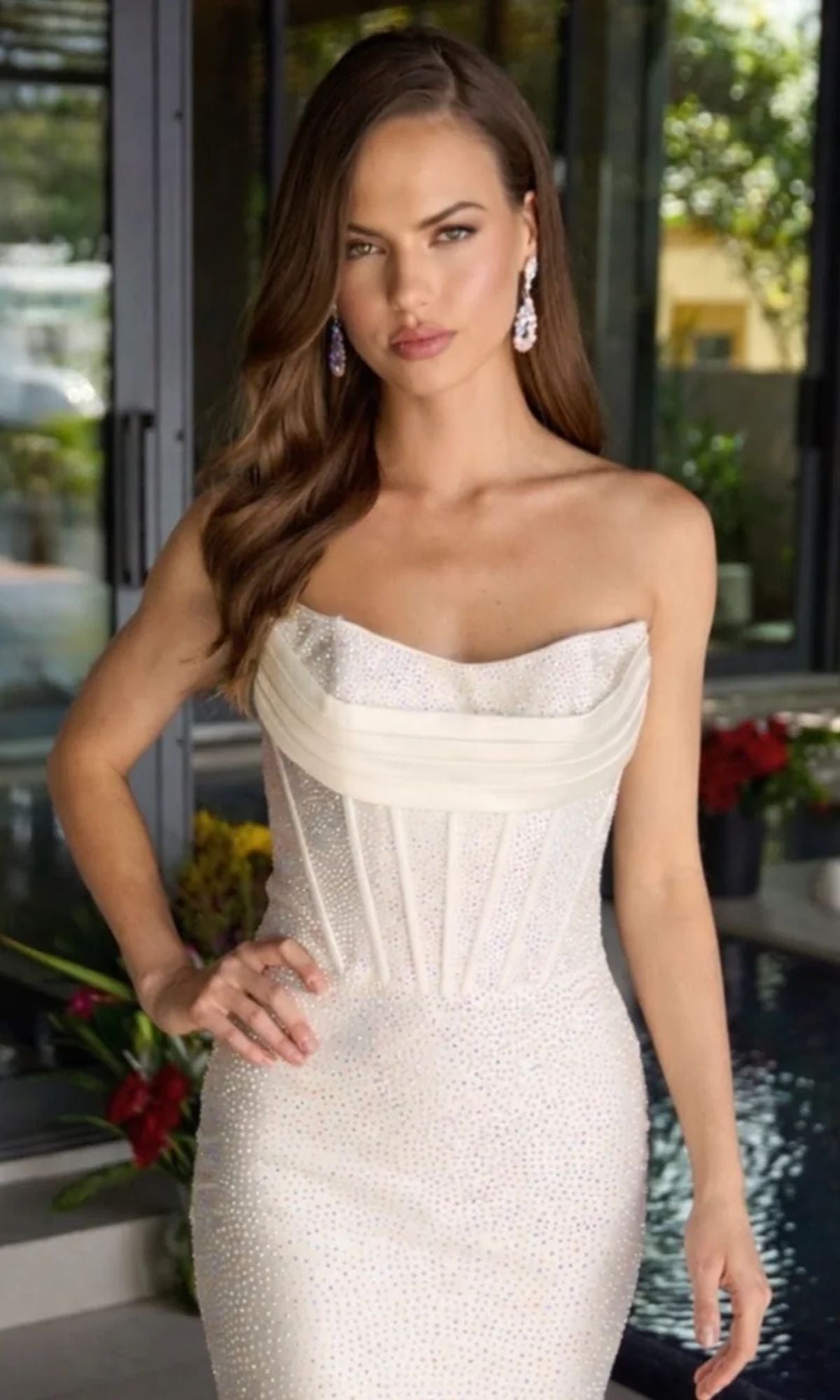 A woman with long brown hair, wearing the Ava Presley 47302 short homecoming dress—featuring a strapless white design with a corset bodice and crystal embellishments—stands indoors near a window and pool, looking directly at the camera.
