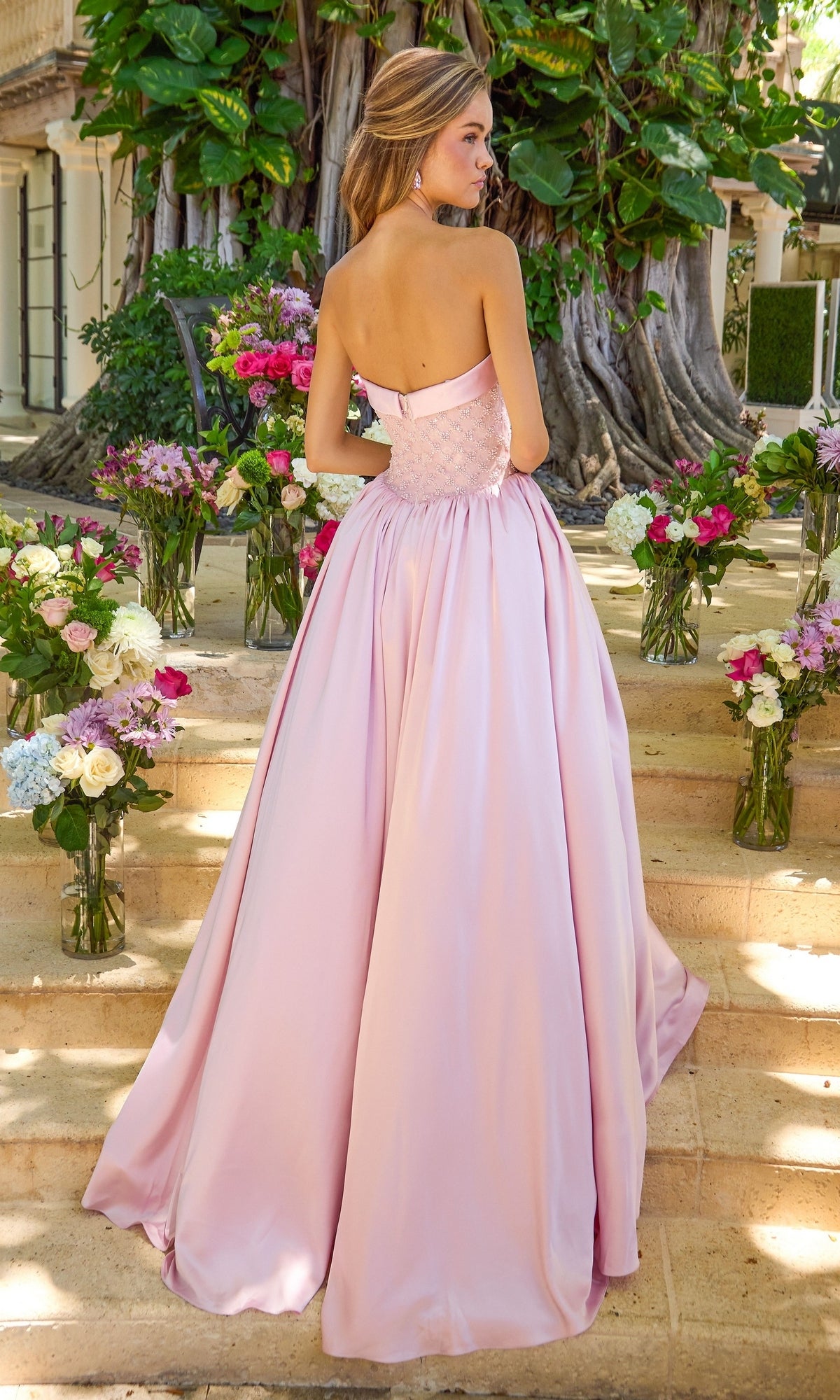 A woman stands on stone steps wearing a light pink, strapless, beaded bodice Ava Presley 47363 ball gown, surrounded by vases of flowers.