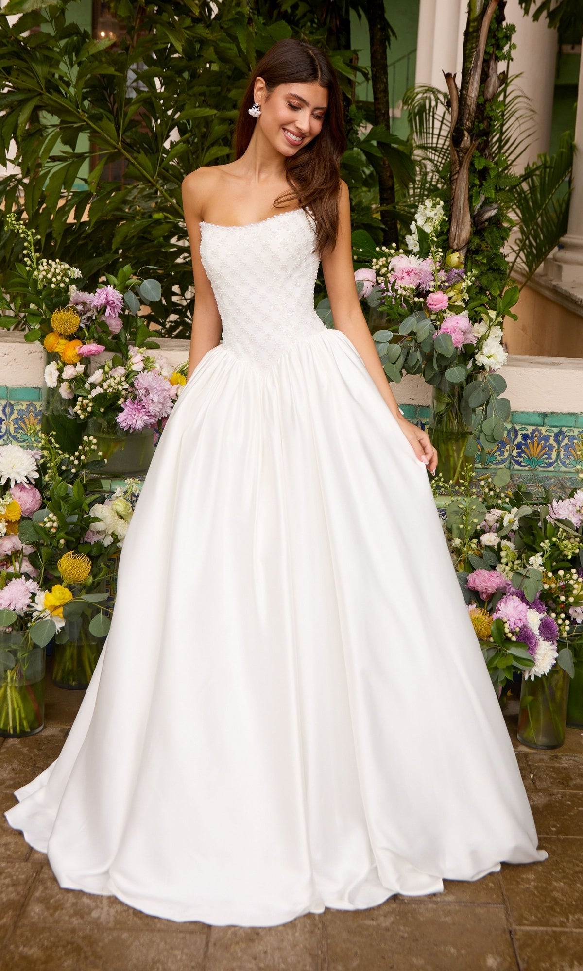A person wears a strapless, floor-length white Ava Presley 47364 ball gown with a textured bodice and full skirt, standing in front of floral arrangements.