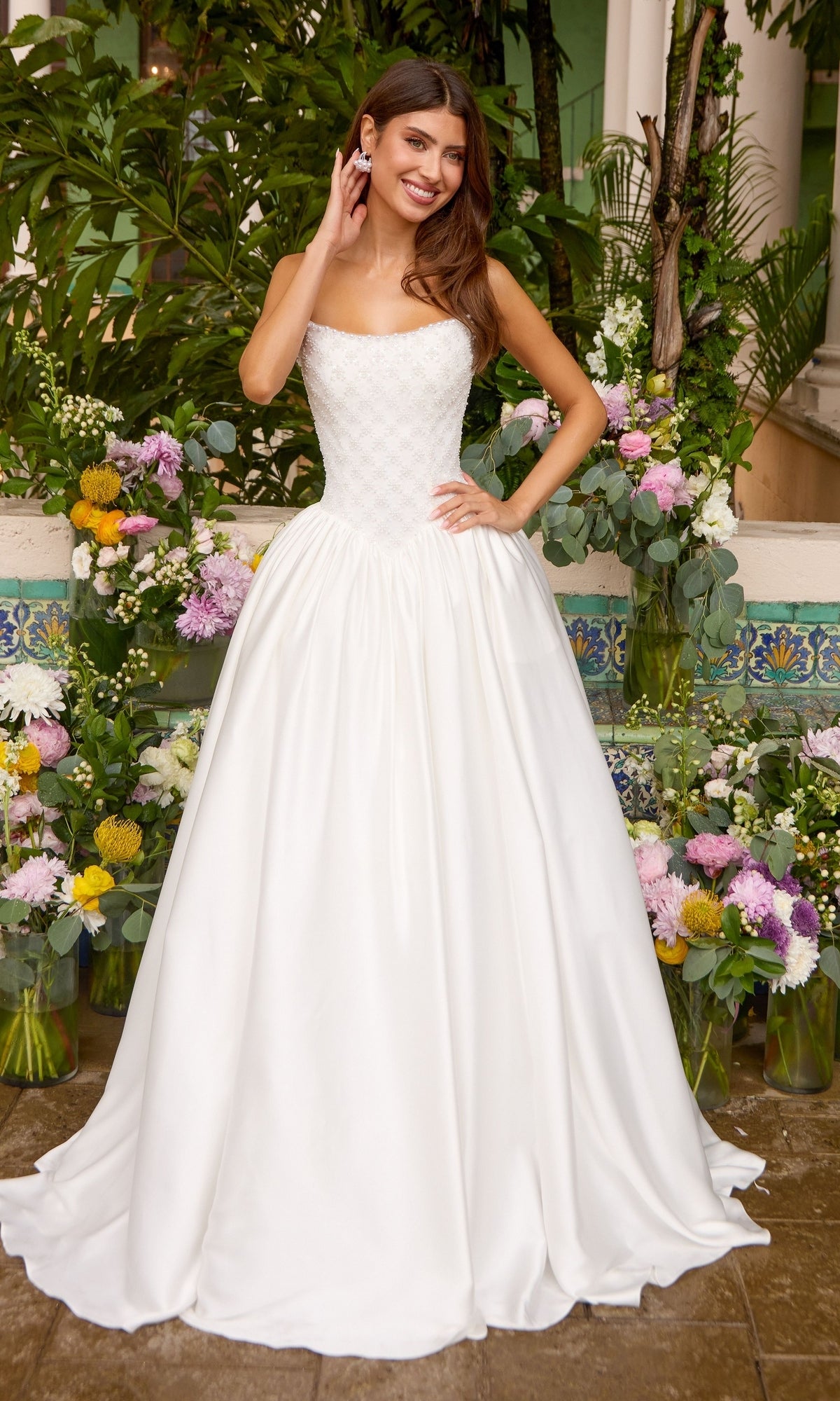 A woman wears an Ava Presley 47364 strapless, floor-length white gown with a beaded bodice and a full, smooth skirt, standing in front of floral arrangements.