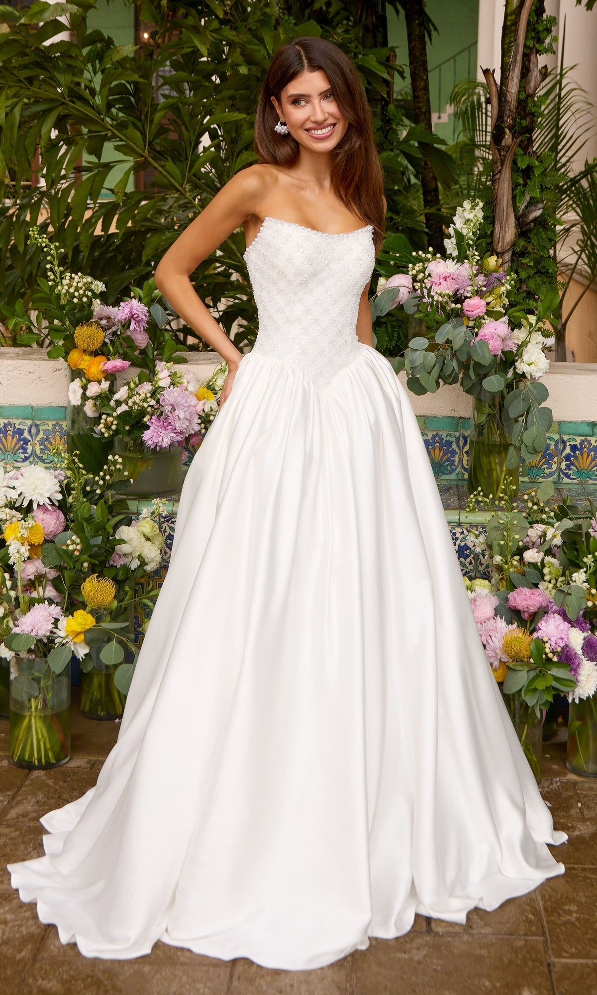 Strapless floor-length Ava Presley 47364 ball gown featuring a beaded bodice and a full skirt, posed before arrangements of assorted flowers.
