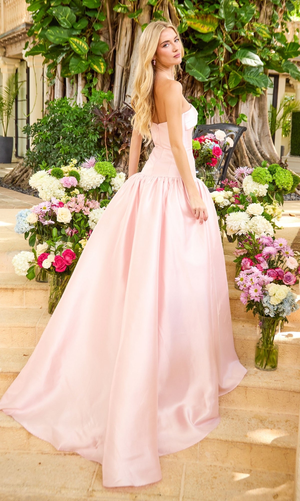 A woman wearing a minimalist Ava Presley 47374 prom ball gown stands on stone steps surrounded by vases of flowers.