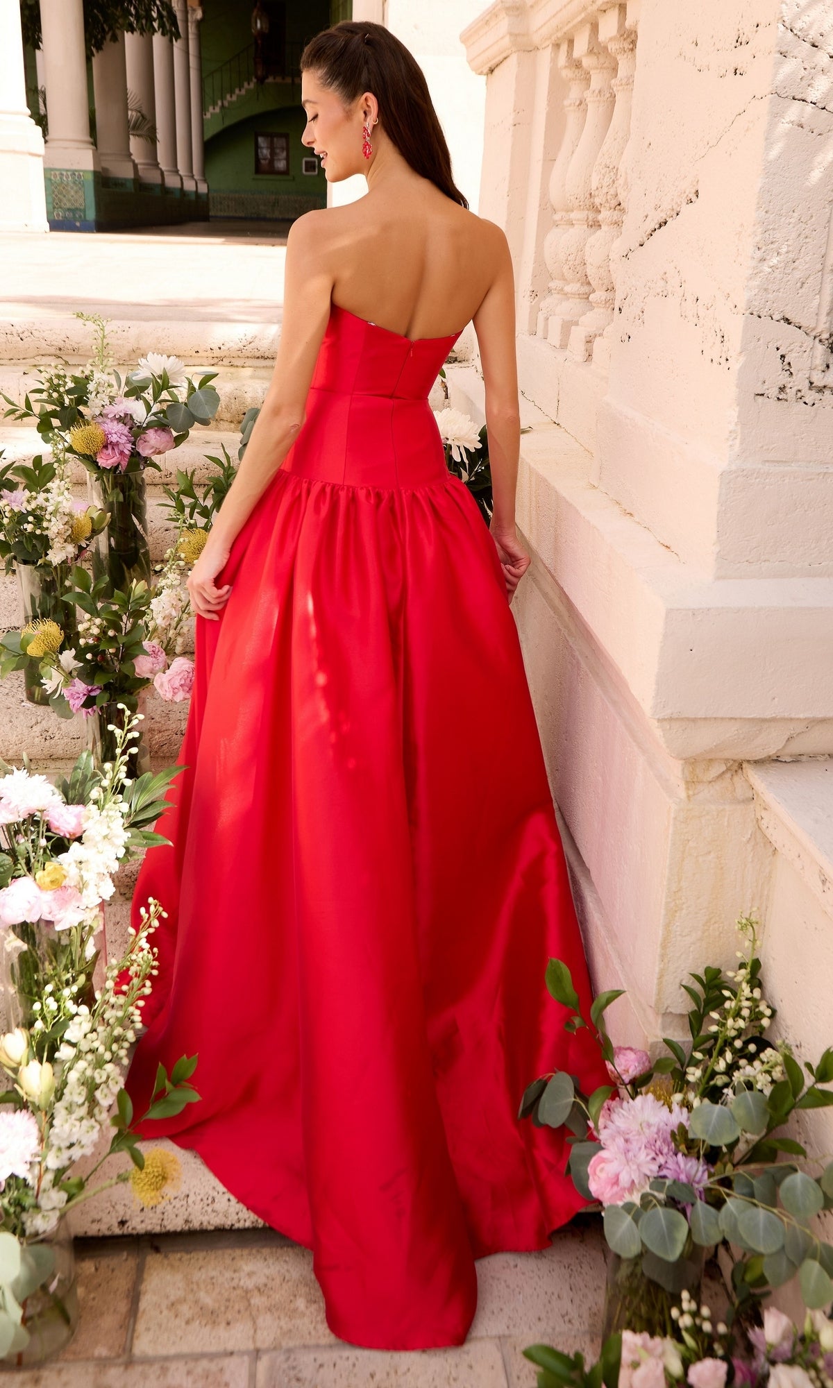 A person wearing a minimalist Ava Presley 47374 prom ball gown, standing on stone steps lined with flower arrangements.