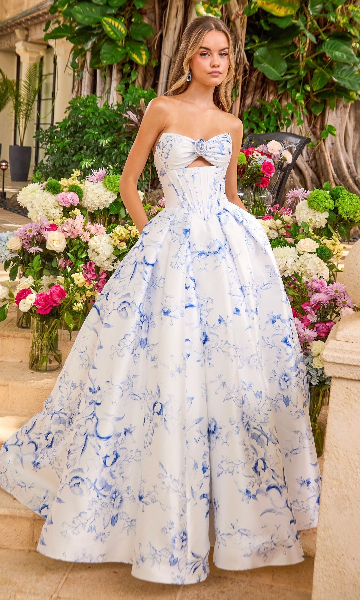 A woman wears a strapless, white and blue floral Ava Presley 47377 floor-length prom dress with a bodice cutout, posed outdoors among vases of flowers.