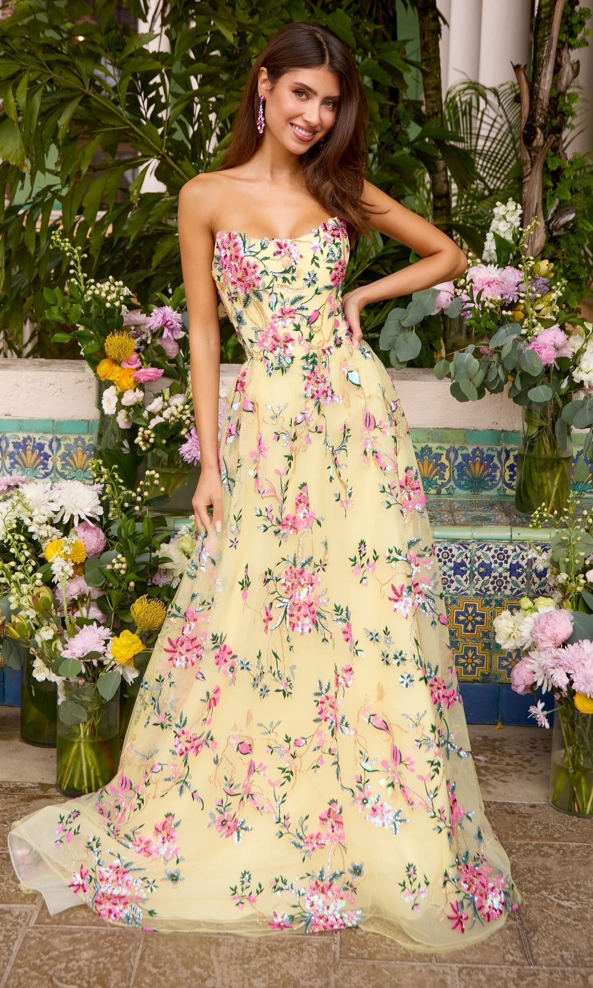 A woman wears an Ava Presley 47391 strapless, floor-length yellow prom dress featuring pink and green floral embroidery.
