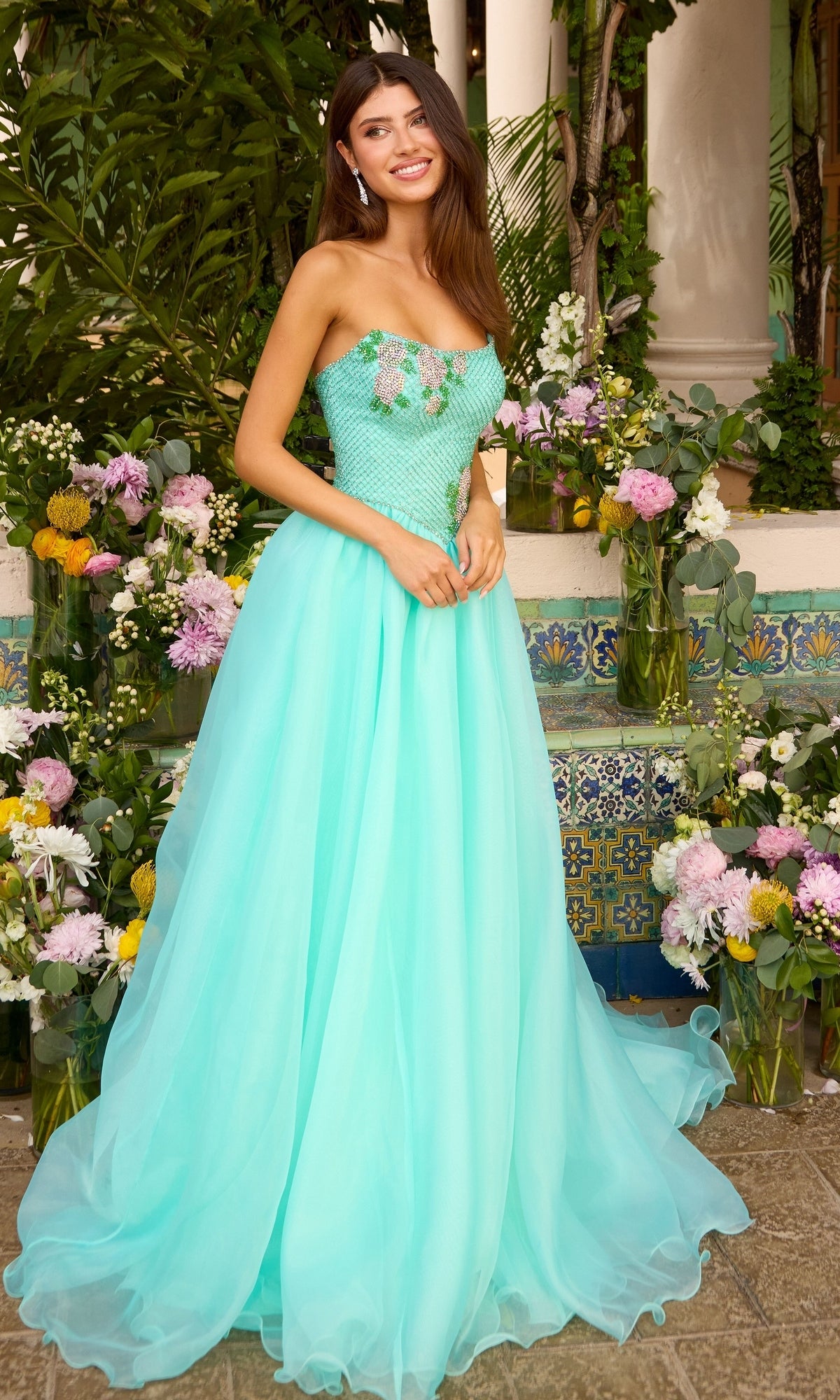 A woman wearing a turquoise strapless Long Prom Dress: Ava Presley 47424 with floral bodice embellishments, standing among flower arrangements.