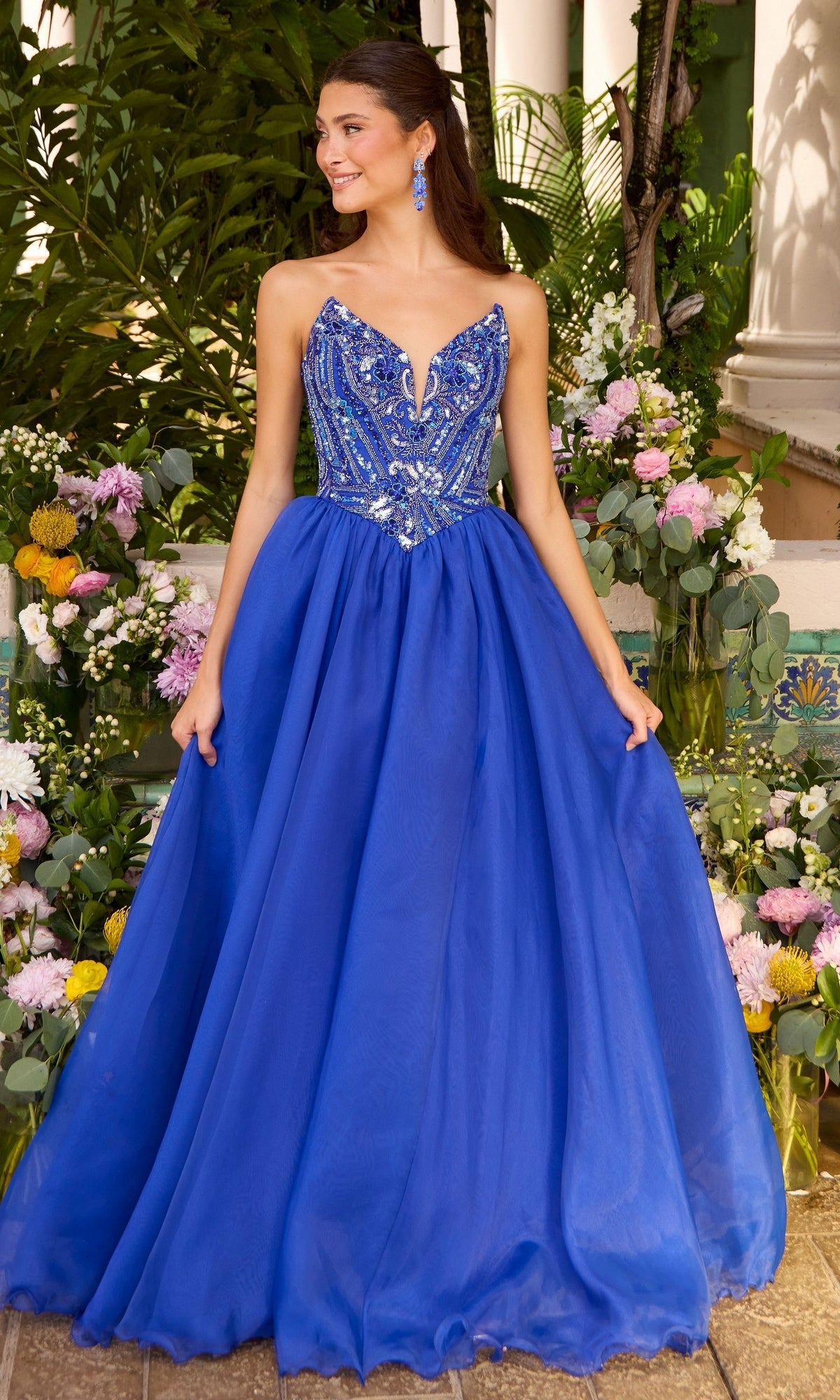 A woman wearing a royal blue Ava Presley 47430 ball gown with a beaded, plunging neckline and a full tulle skirt, standing in front of floral arrangements.