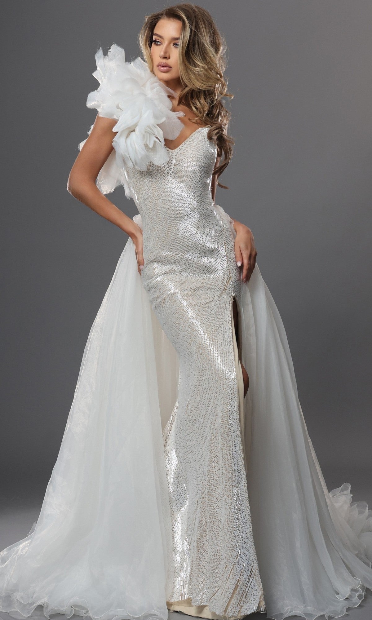A woman in the Long Formal Dress: Jovani 47895 in off white, featuring a thigh-high slit, a prominent white floral shoulder detail, and a flowing train, stands against a gray background.