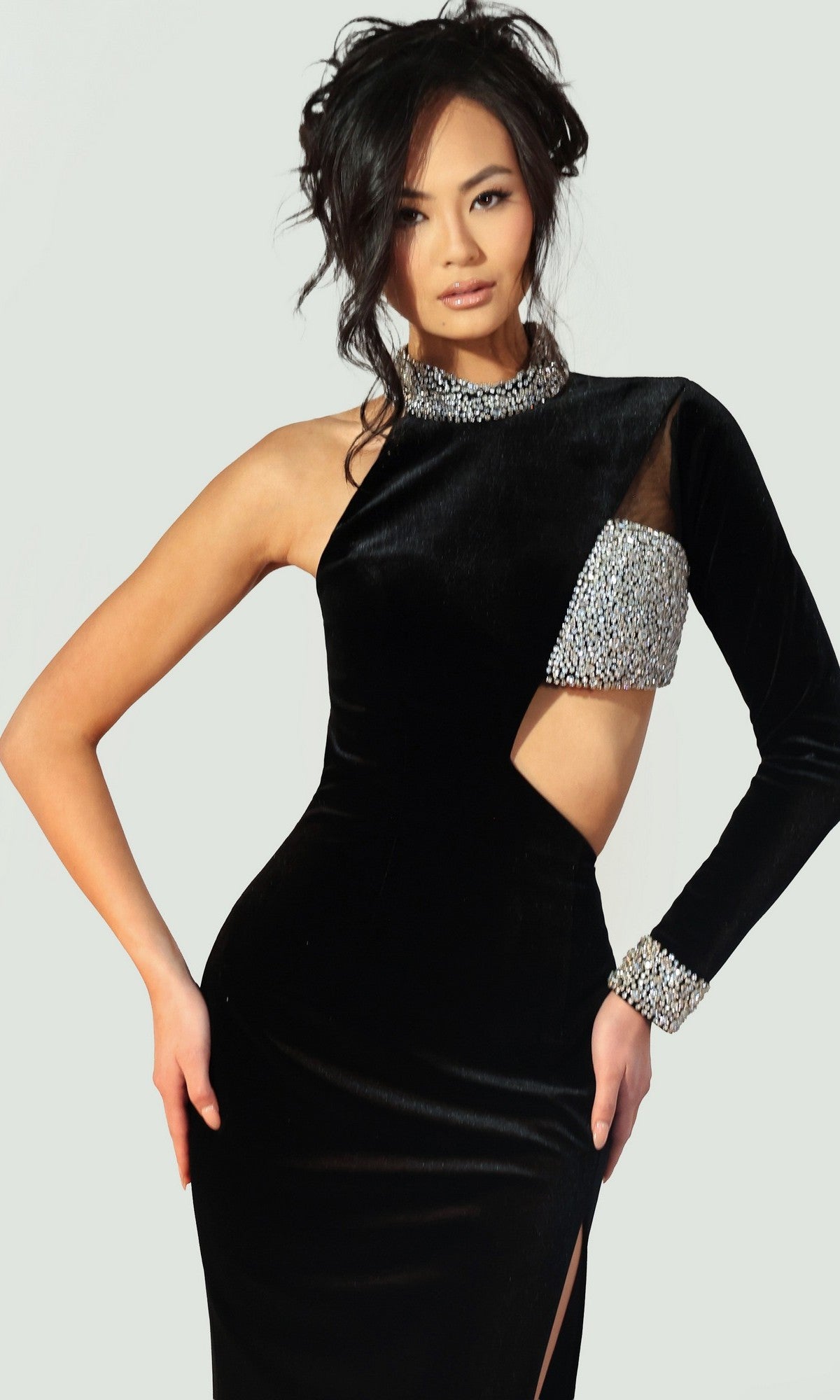 A woman poses against a plain light background wearing the Long Prom Dress: Jovani 48481, a black gown with an asymmetrical design, one long sleeve, silver embellishments, and a side cutout.