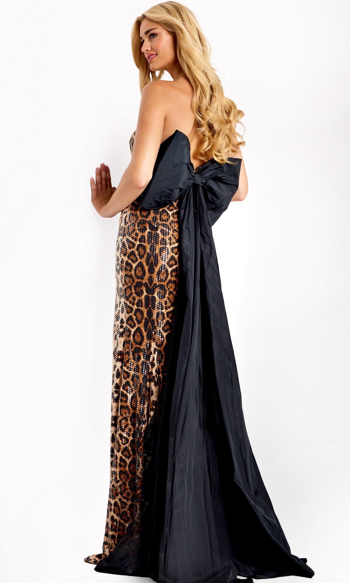 A woman stands sideways against a white background wearing the Jovani 48651 long prom dress—a strapless, leopard-print sequin gown featuring a sweetheart neckline, large black bow, and long black train.