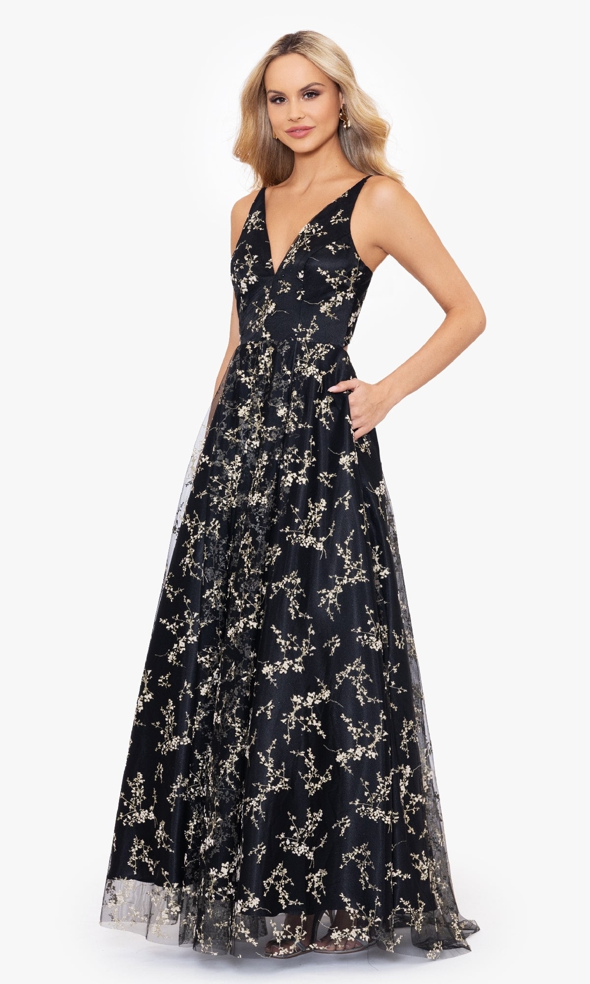 A woman poses against a plain white background wearing the Blondie Nites 4866BN Floral-Print Long Prom Dress—a sleeveless black gown with pockets and gold floral embroidery.