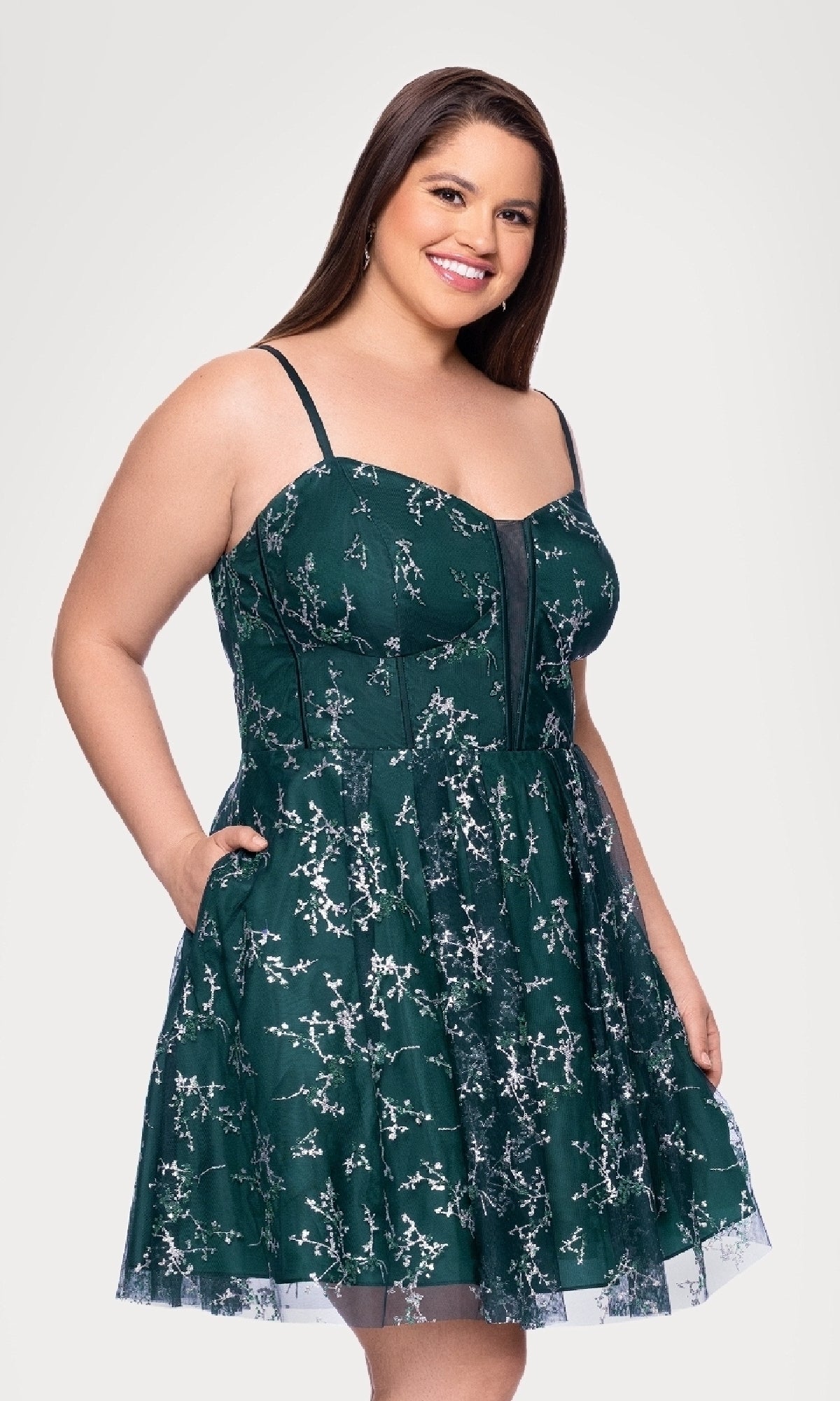A woman models the Plus Short Green Hoco Dress by Blondie Nites 4884BNW—a sleeveless, dark green floral style with thin straps—standing and smiling against a plain white background.