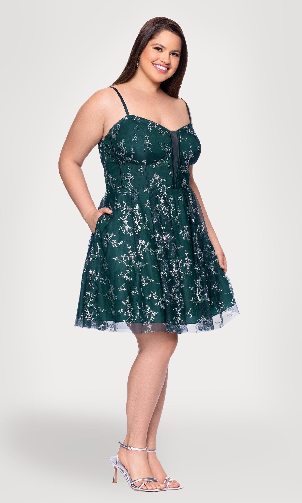 A woman in the Plus Short Green Hoco Dress: Blondie Nites 4884BNW with floral embroidery, thin straps, and pockets pairs it with silver heels, standing against a plain light background.