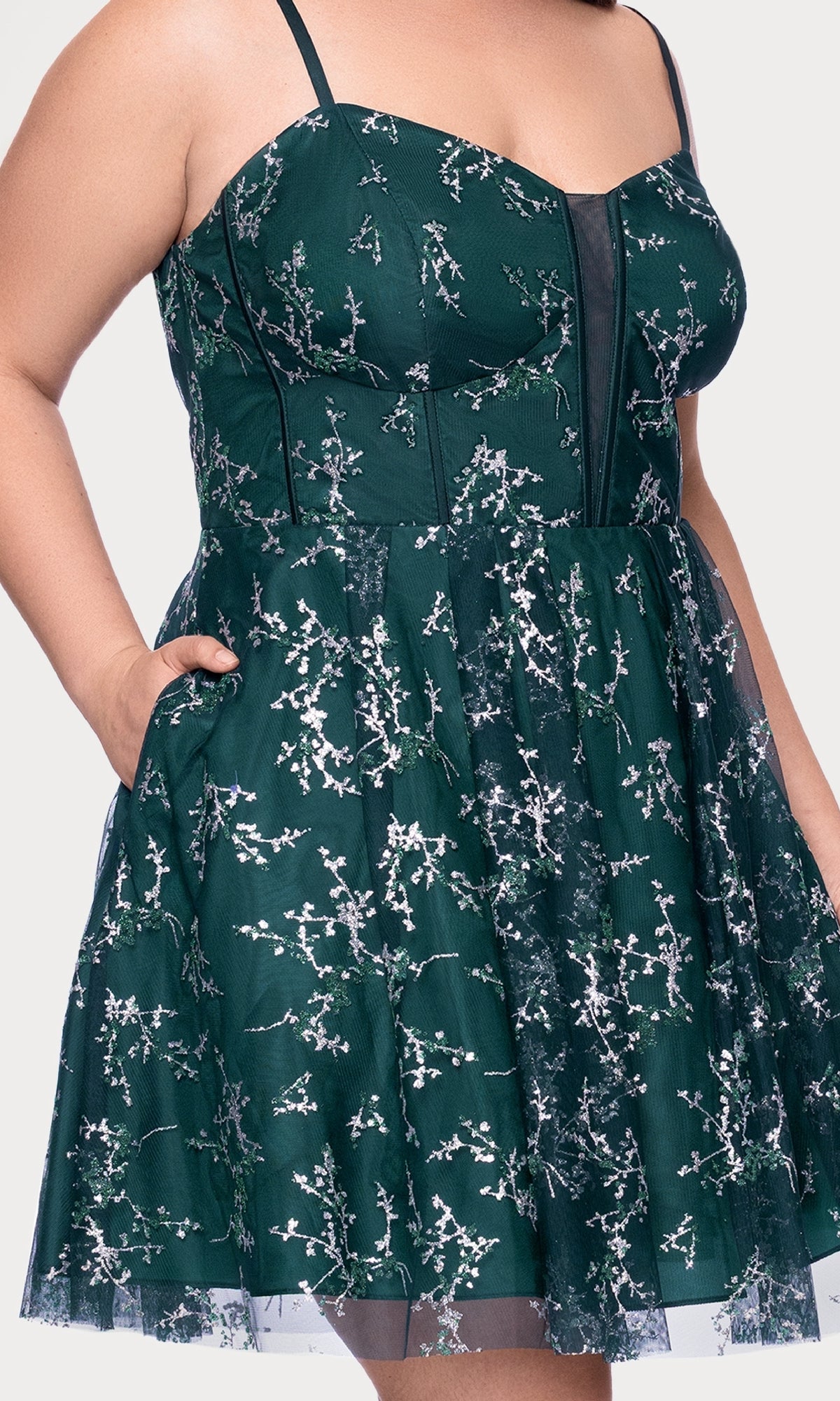 Someone wearing the Plus Short Green Hoco Dress: Blondie Nites 4884BNW—a sleeveless dark green plus-size homecoming dress with silver floral embroidery, mesh accents, a fitted bodice, and flared skirt.