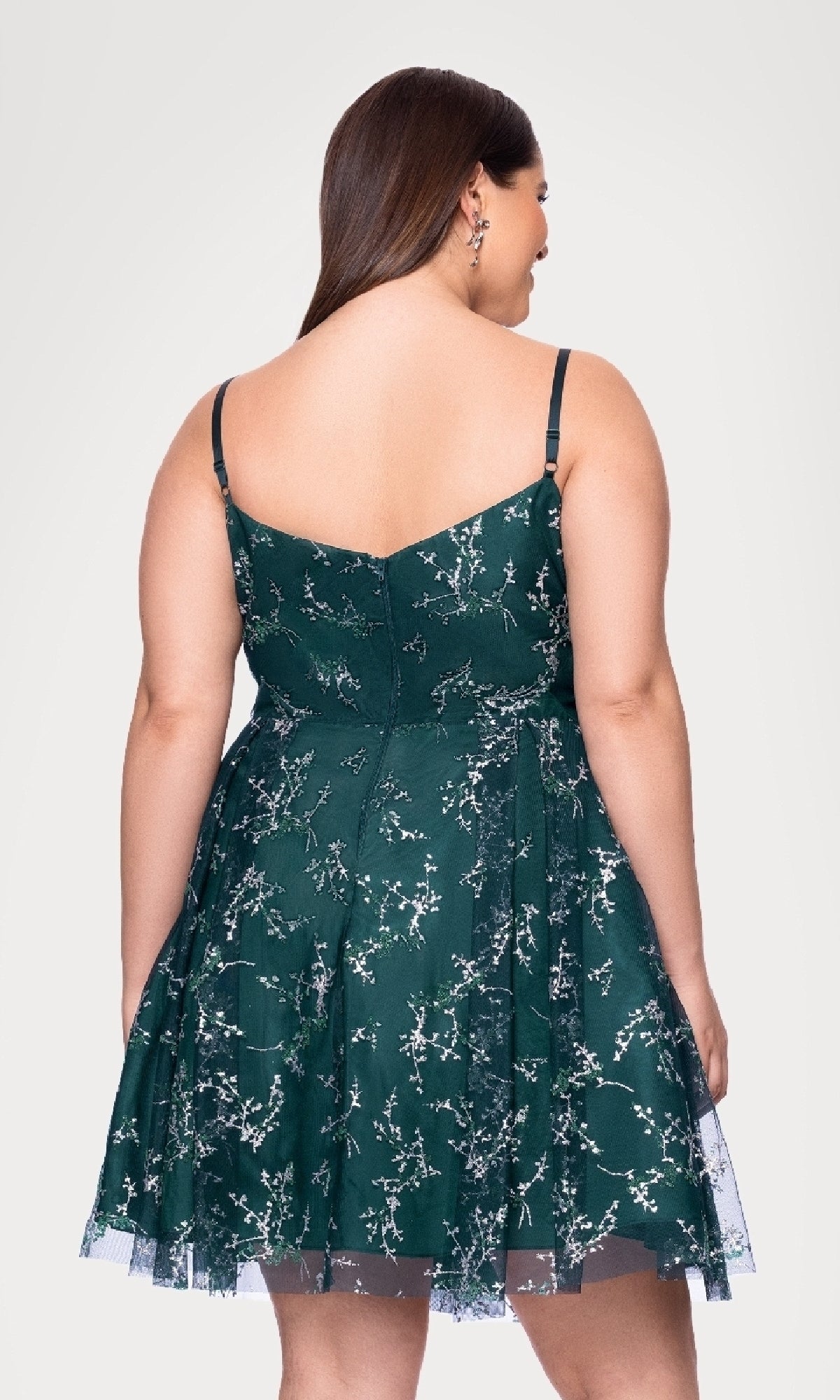 A woman models the Plus Short Green Hoco Dress: Blondie Nites 4884BNW, featuring a dark floral sleeveless design with thin straps, photographed from the back against a white background.