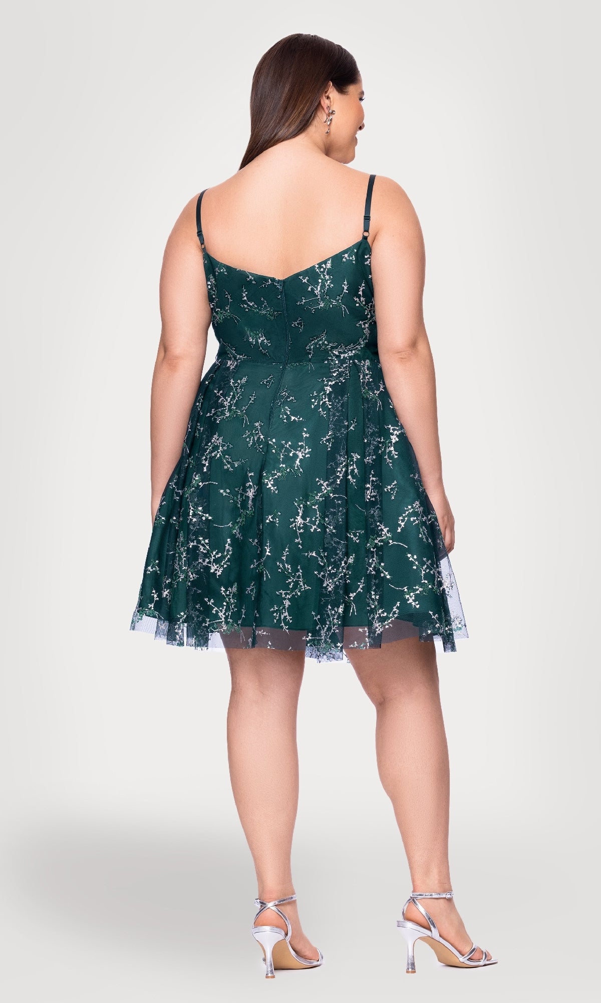A woman models the Plus Short Green Hoco Dress: Blondie Nites 4884BNW, a green sleeveless cocktail dress with floral lace and spaghetti straps, paired with silver high-heeled sandals, photographed from the back against a plain background.