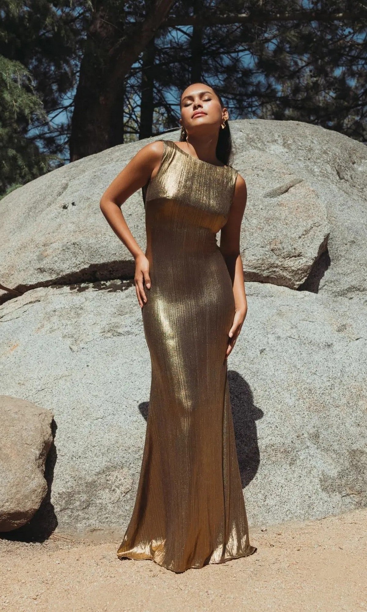 A woman stands outdoors on sandy ground before large rocks and trees, wearing the V-Back Long Metallic Prom Dress: Mac Duggal 49087.