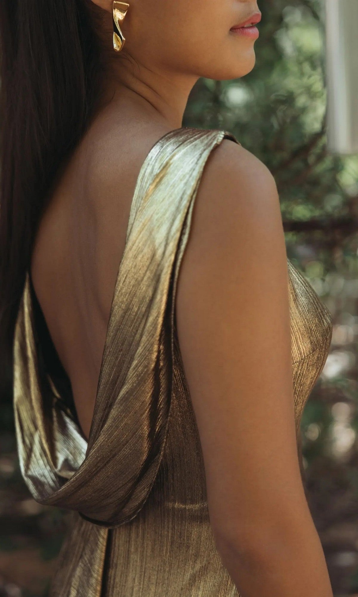 A woman stands outdoors in the Mac Duggal 49087 V-Back Long Metallic Prom Dress, featuring a gold metallic finish and draped back, paired with gold earrings as she faces to the side.