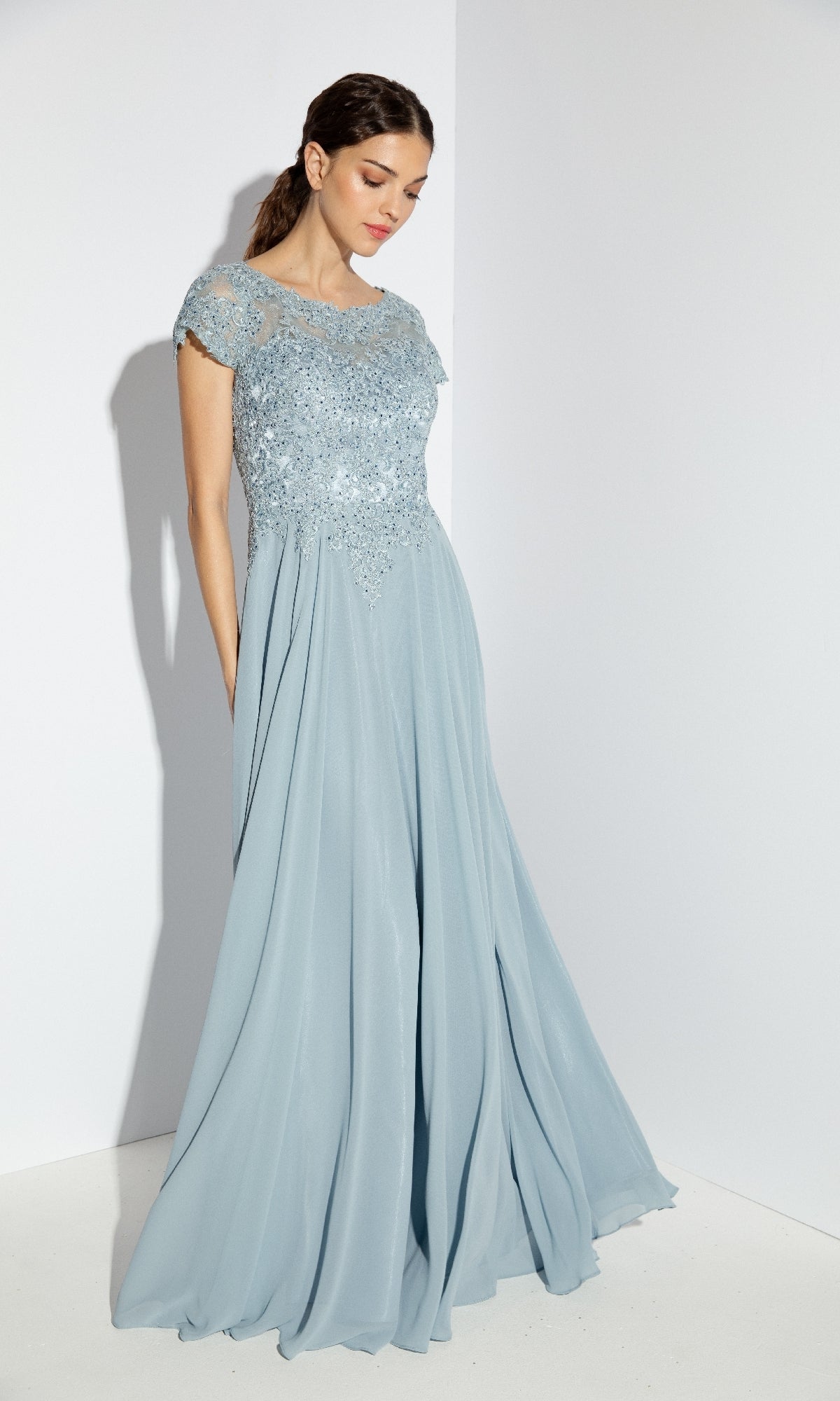 A woman stands against a white background wearing the Eureka 4909, a light blue short-sleeve long A-line formal dress with an embroidered lace bodice and flowing chiffon skirt, styled elegantly for dance.