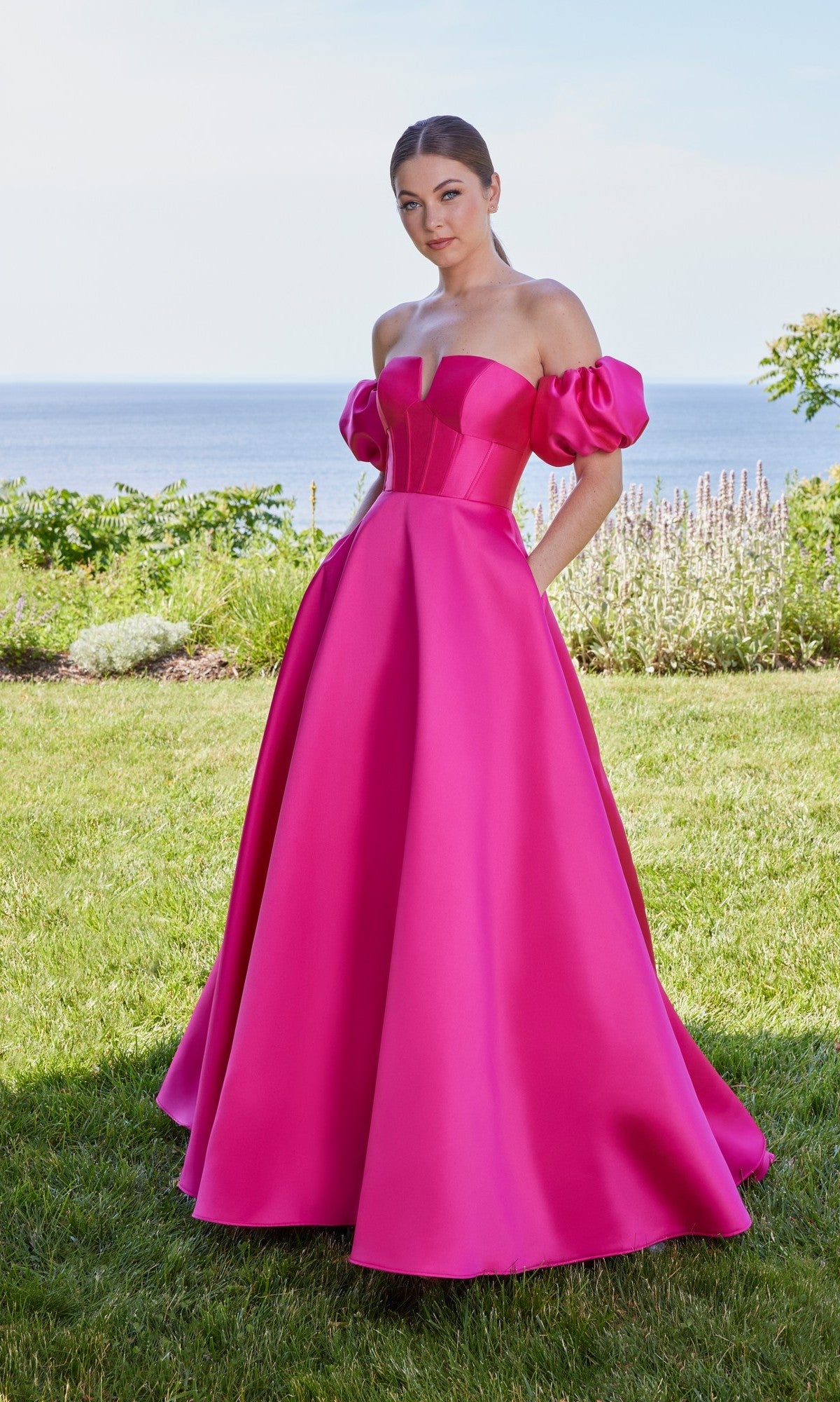 A woman stands on the grass by the ocean wearing the Morilee 49129 Long Prom Dress in fuchsia, featuring off-the-shoulder puffed sleeves and a corset bodice.