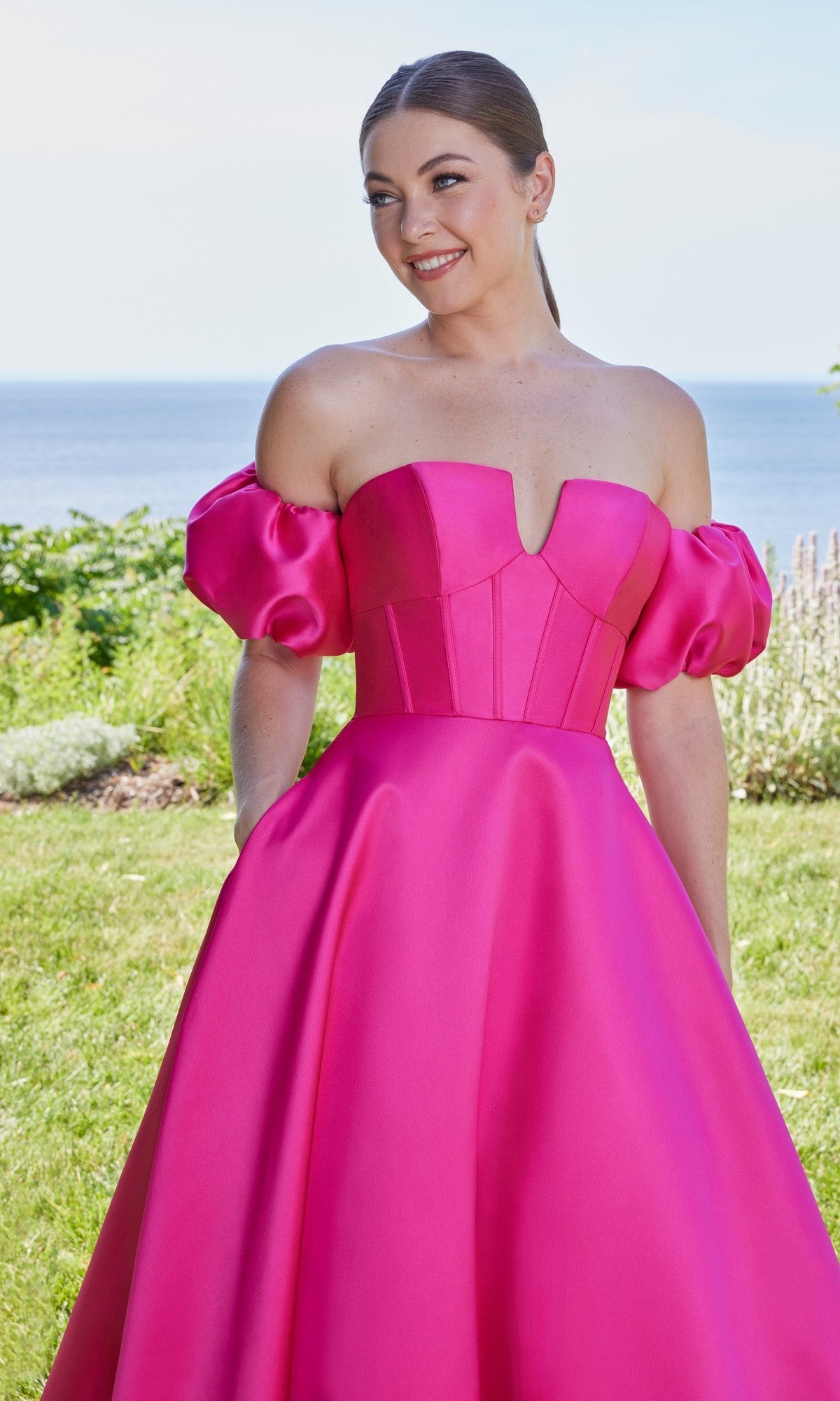 A woman stands outdoors by the water, wearing the Morilee 49129 long prom dress featuring bright pink fabric, puffed sleeves, and a fitted corset bodice.
