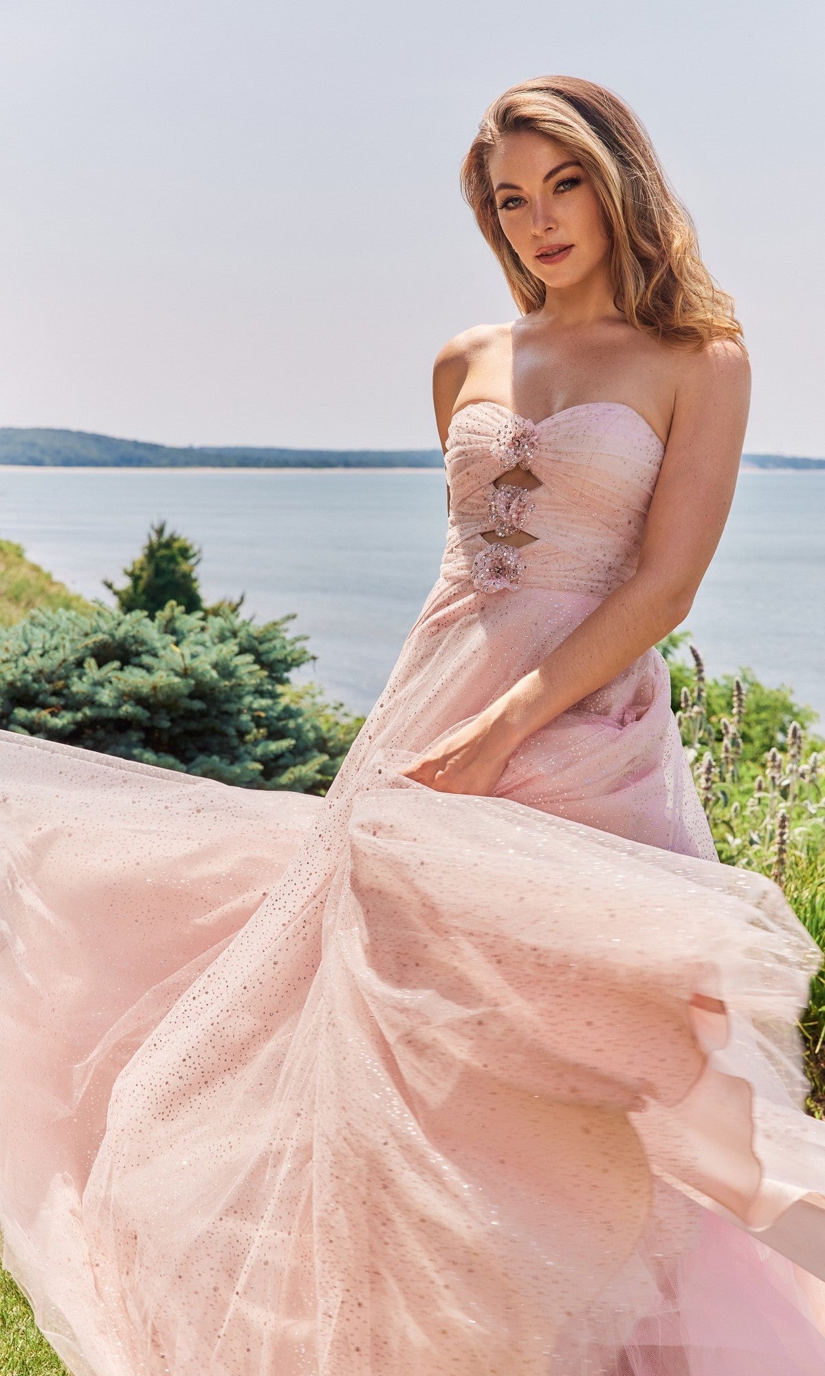 A woman in a blush Morilee 49137 long prom dress with a strapless bodice and elegant a-line skirt stands outdoors by the water, holding her gown, with greenery and shoreline in the background.