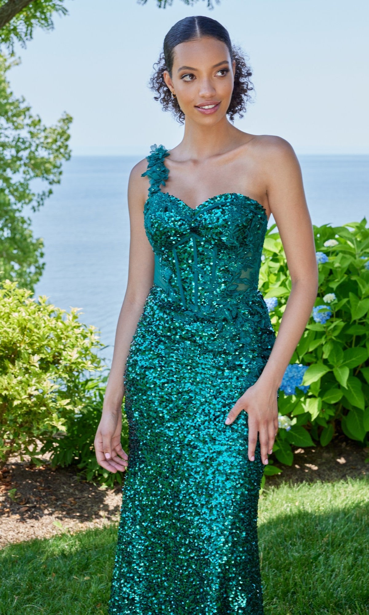 A woman stands outdoors wearing the Morilee 49163 long prom dress in emerald, featuring a one-shoulder design with sequins and floral details, a fitted bodice, and nature’s greenery and water in the background.