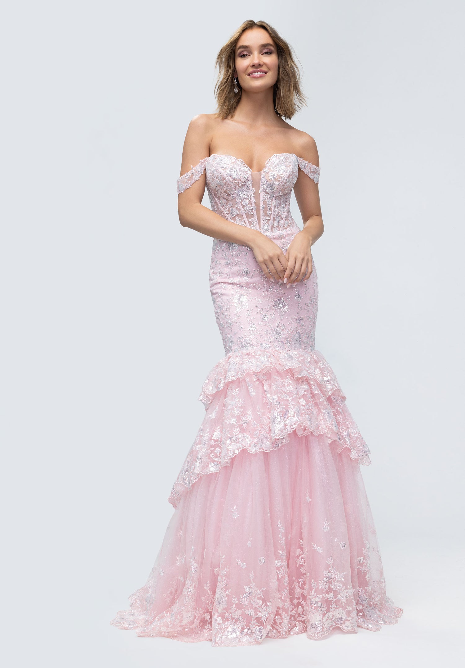 A woman wears the Morilee 49274 long prom dress in blush, featuring an off-shoulder lace corset and layered tulle mermaid skirt, standing against a plain light background.